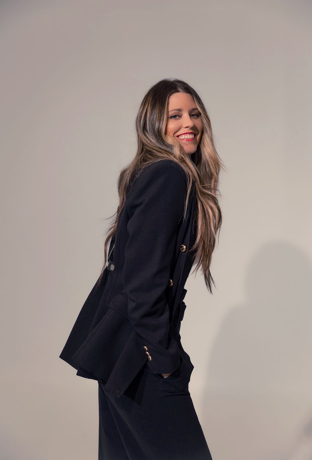 Woman with long hair smiling, wearing a dark blazer, standing against a plain light background.