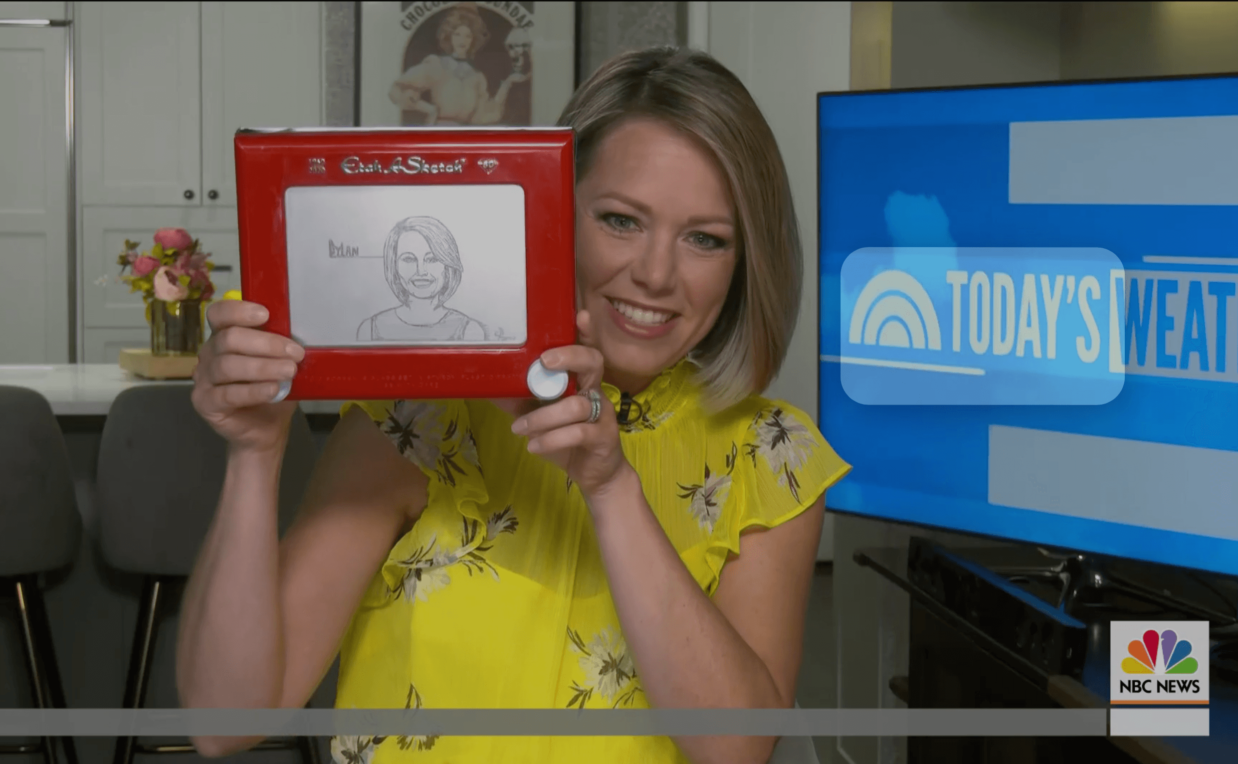 Woman in yellow dress holding a red drawing board with a sketch, smiling, in a room with a TV screen and flowers