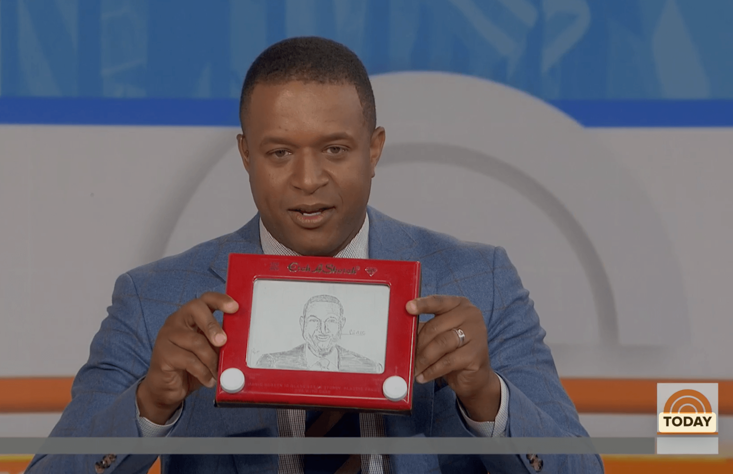 Man in a suit holds a red drawing board with a sketch of a person's face, in a studio setting.