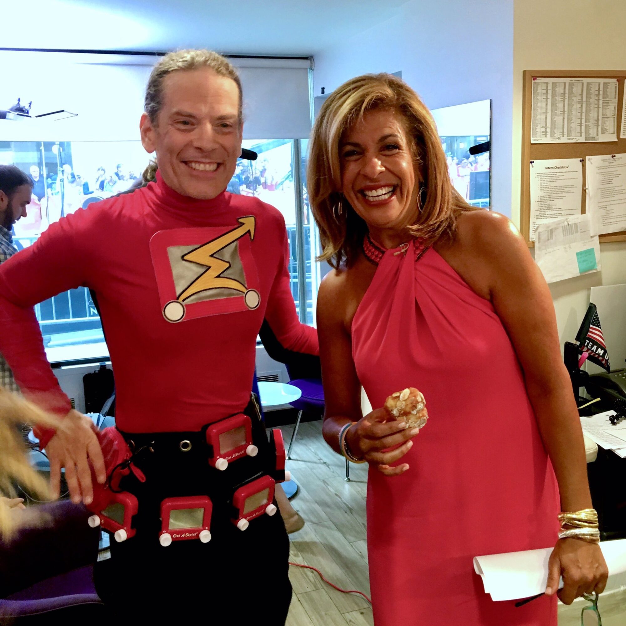 Two smiling women stand indoors, one in a pink dress holding a cupcake, the other in a red shirt with a graphic, both smiling.