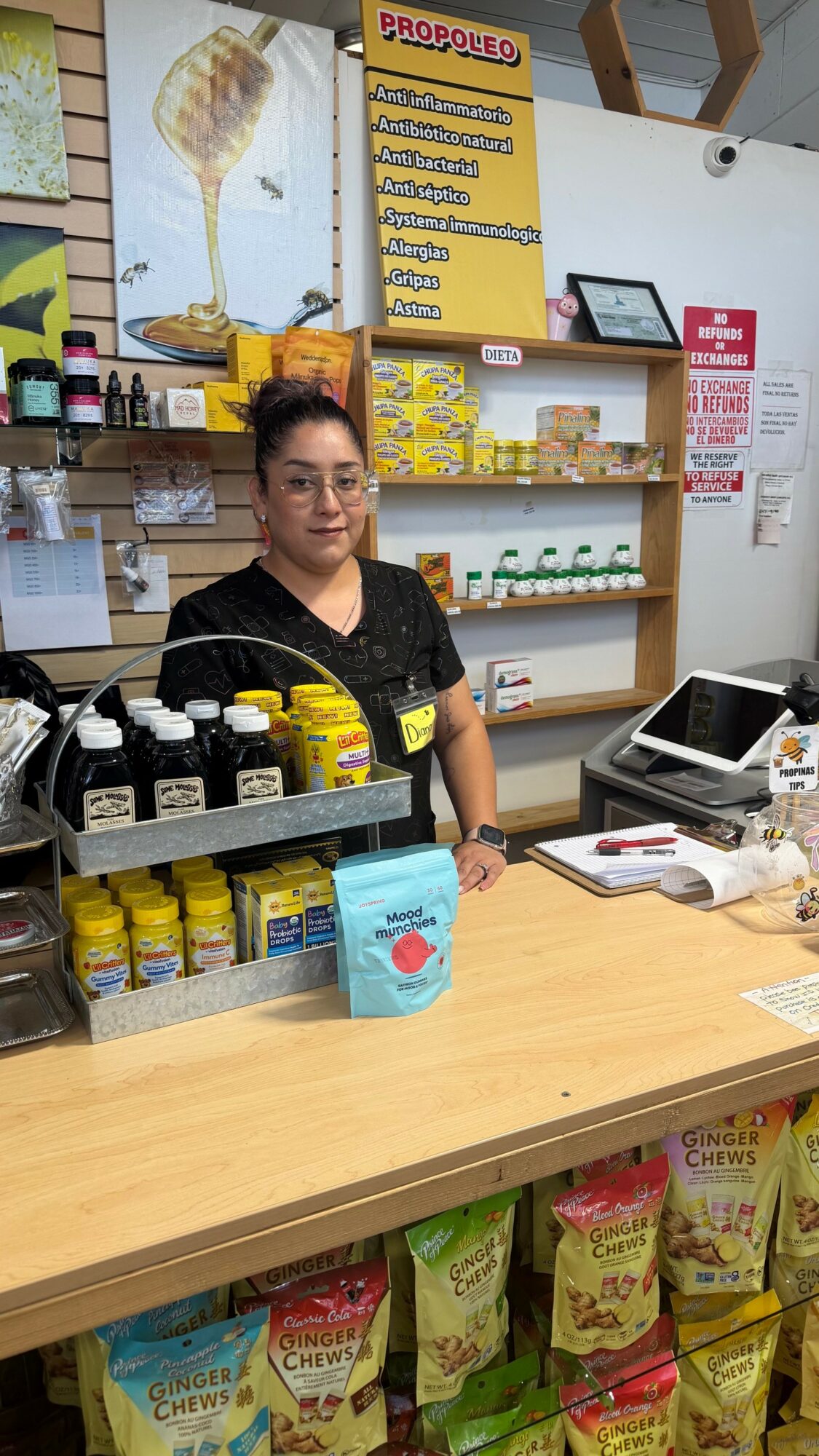 Woman standing behind a store counter with shelves of products and a yellow propolis poster in the background.