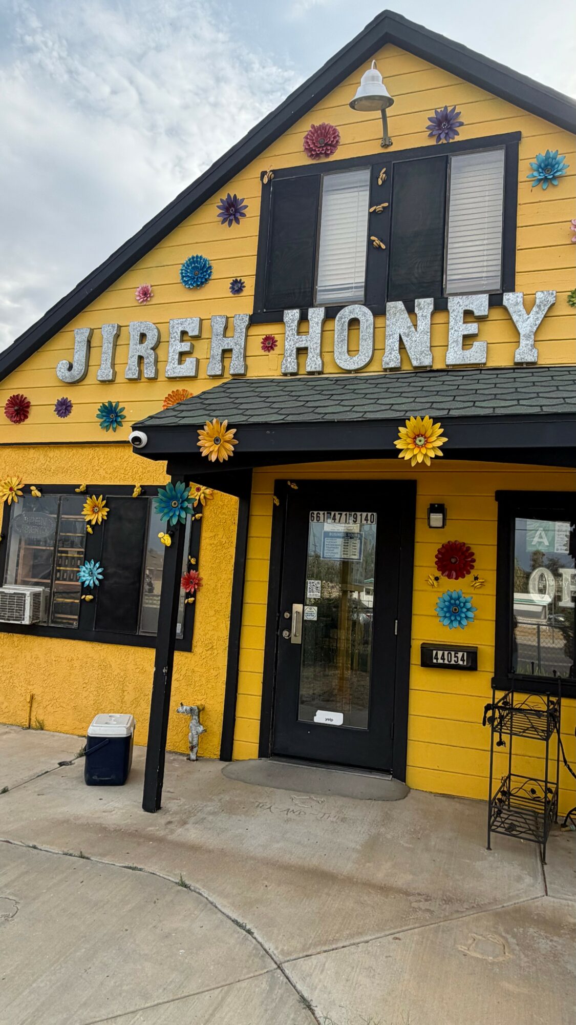 Yellow building with black shutters and colorful flower decorations, sign reads 'Jireh Honey', entrance door with glass window, outdoor seating area.