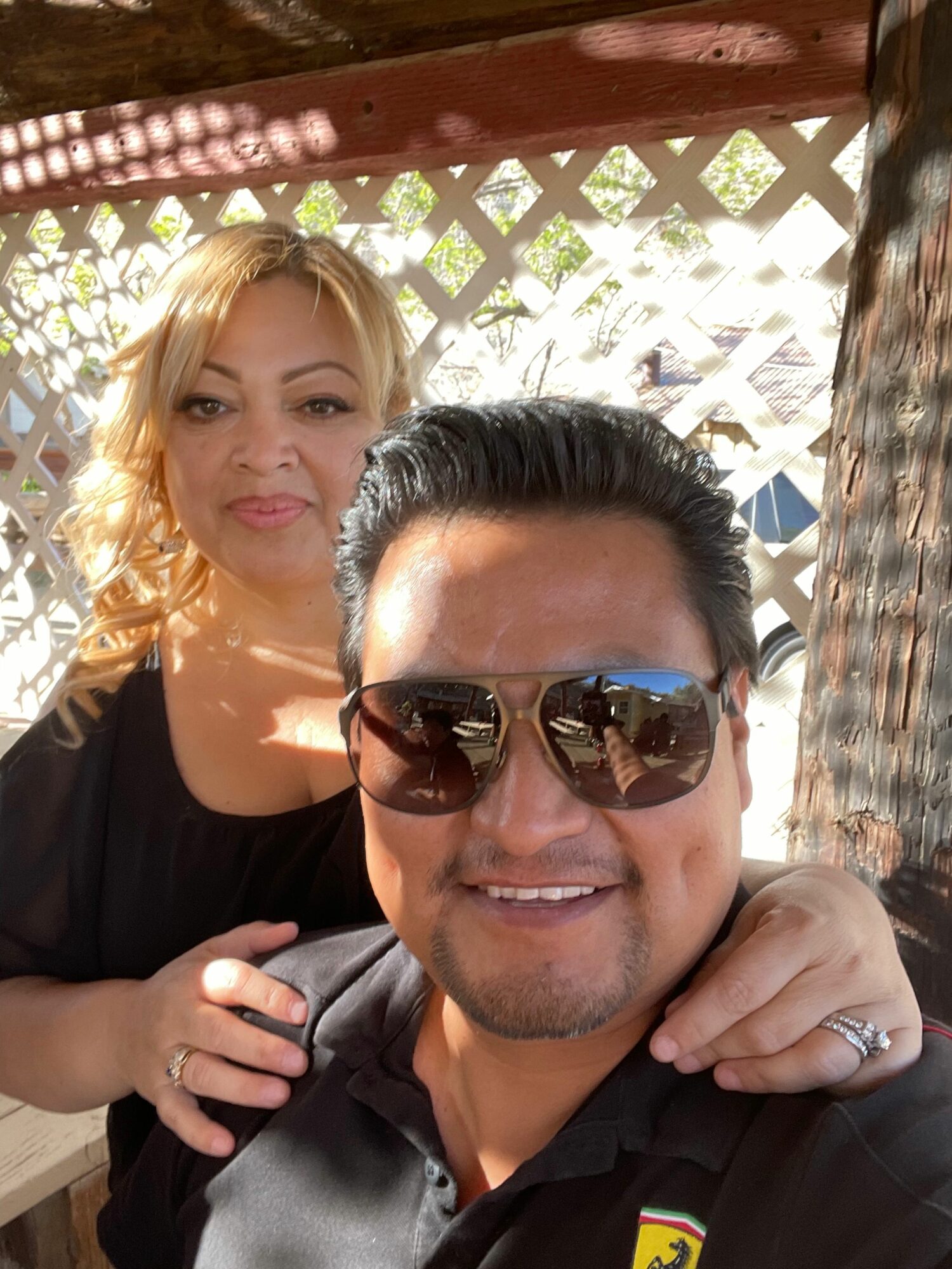 Two people smiling, one wearing sunglasses, outdoors with lattice background, sunlight, and greenery.
