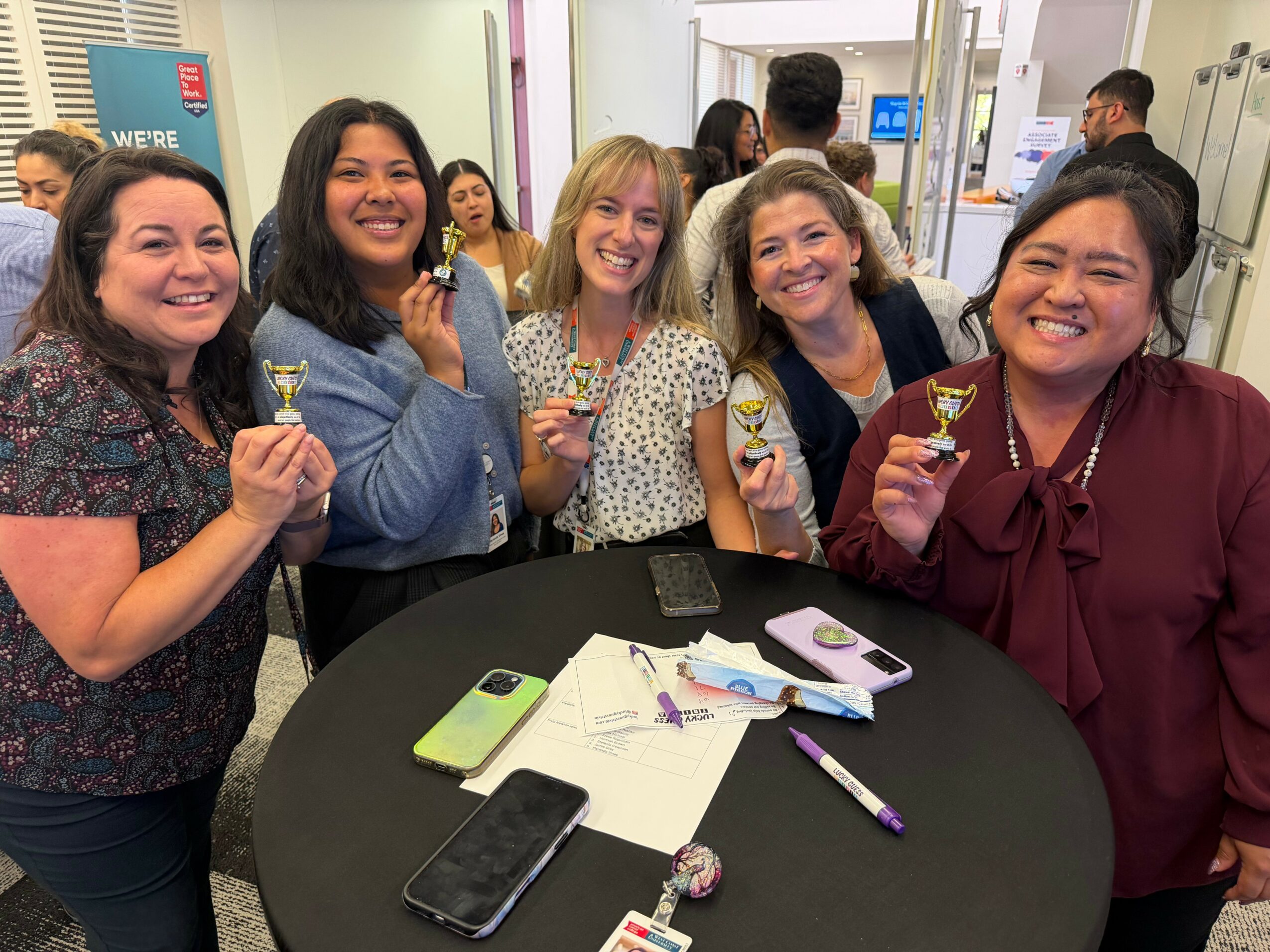 Five women smiling and holding small trophies around a table with papers and phones in a room.