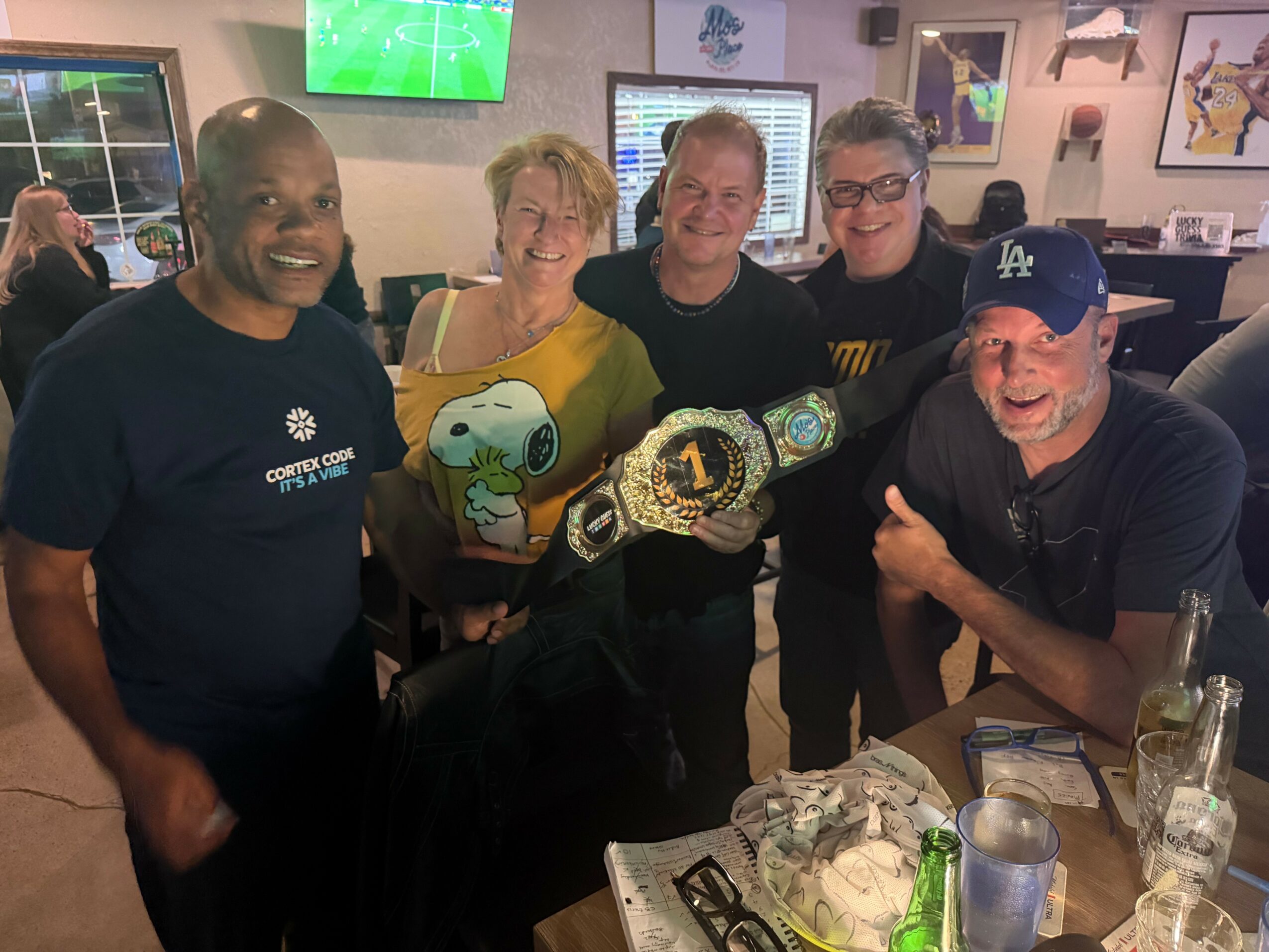 Group of five people smiling indoors, holding a Snoopy plush and championship belts, with drinks on table.