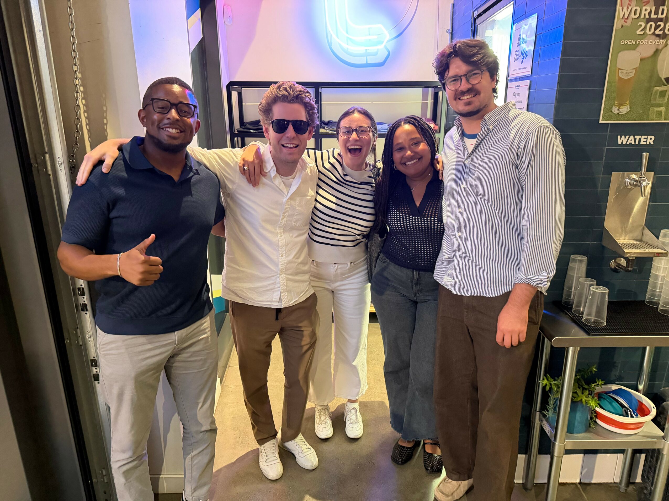Group of five people smiling and standing together indoors, some with arms around each other.