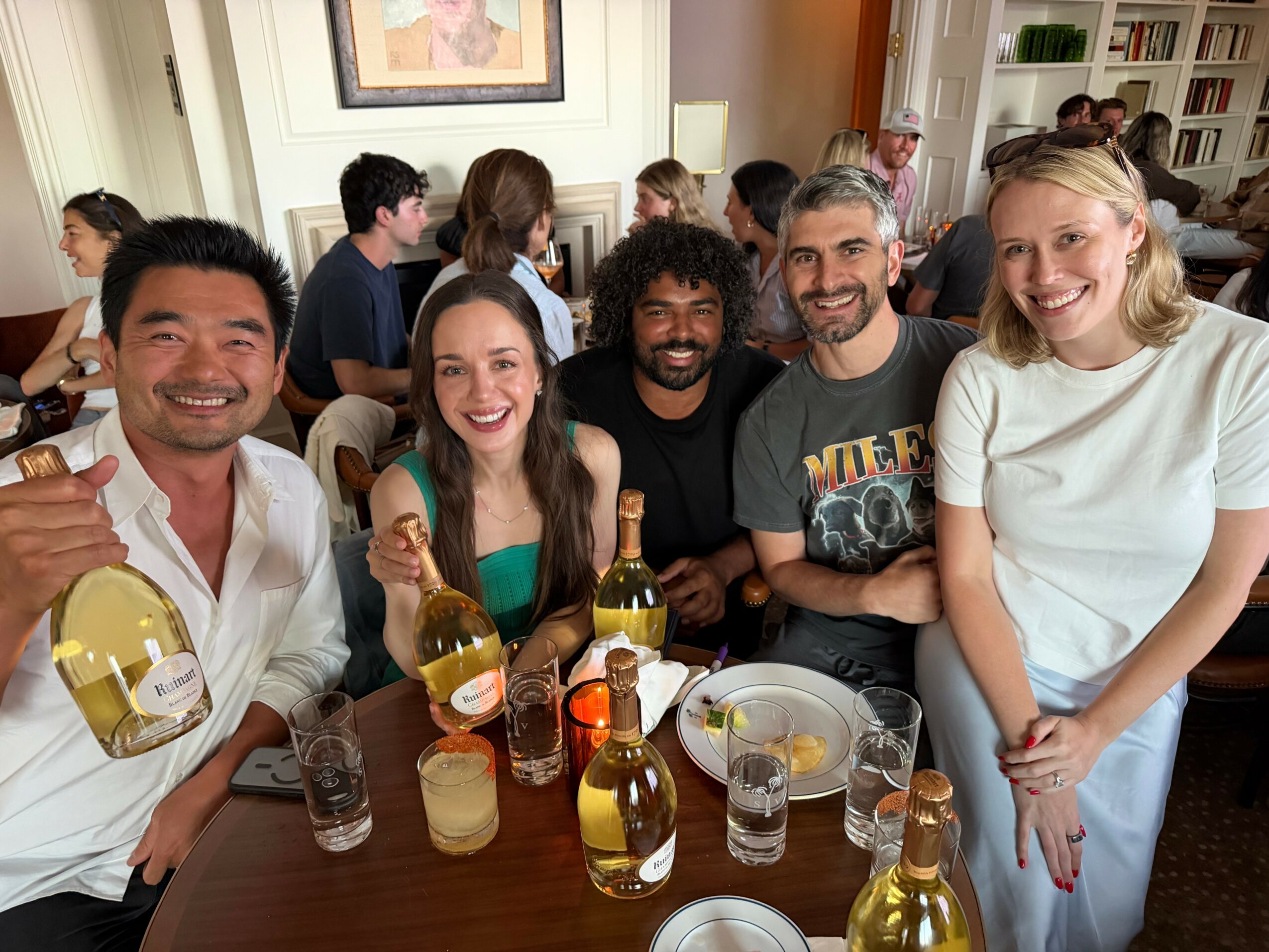 Group of five people smiling at a table with drinks and snacks in a cozy room.