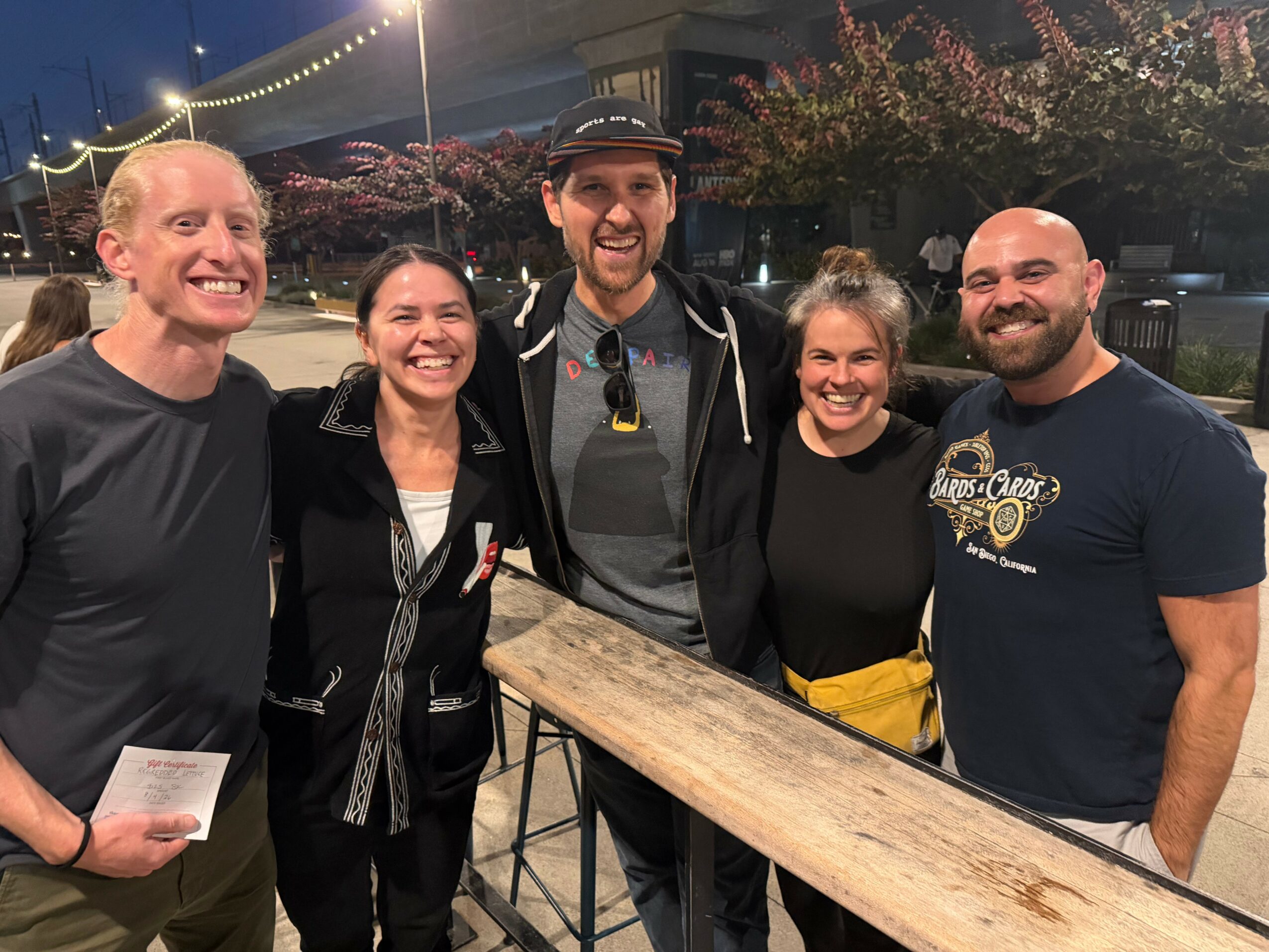 Group of five people smiling outdoors at night, standing behind a wooden table, with string lights overhead.
