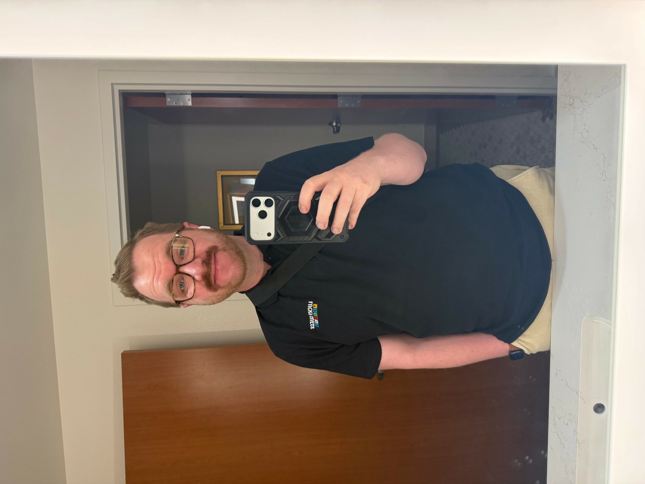 Person with glasses taking a mirror selfie in a room, wearing a black shirt and beige pants, holding a phone with a white case.