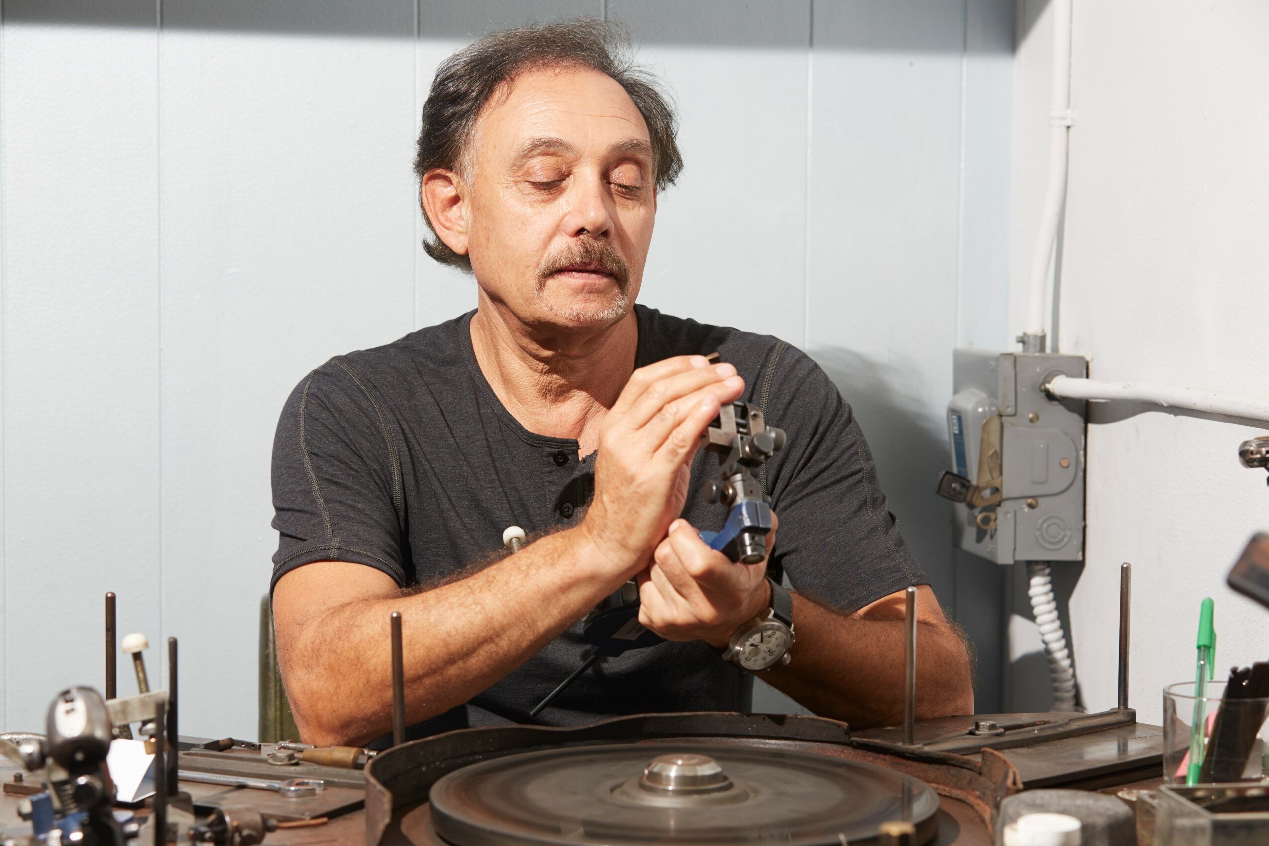 Man working with a small tool on a mechanical part, seated at a workbench in a workshop.