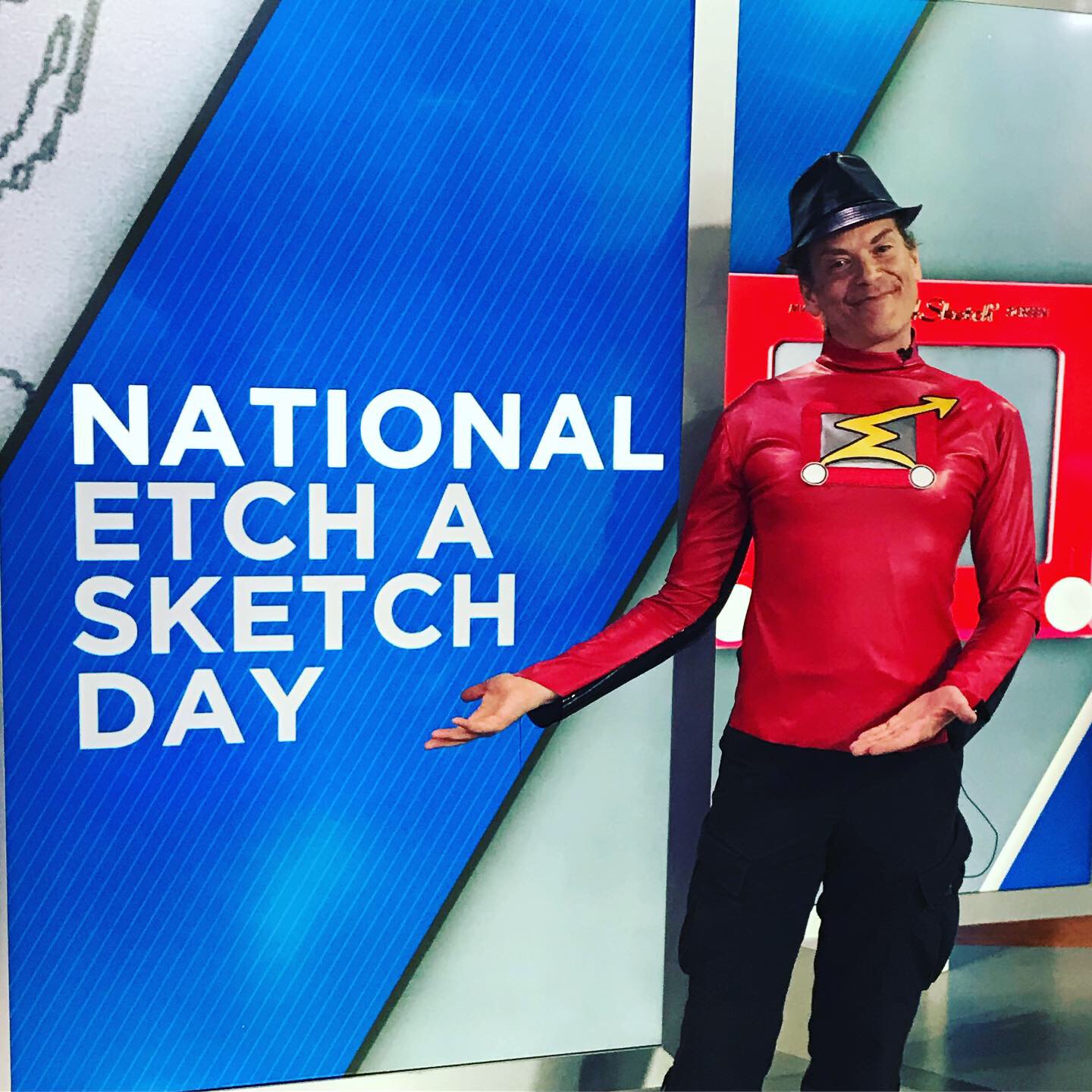 Person dressed as a superhero in a red costume and black hat, standing next to a sign that reads 'National Etch A Sketch Day'.