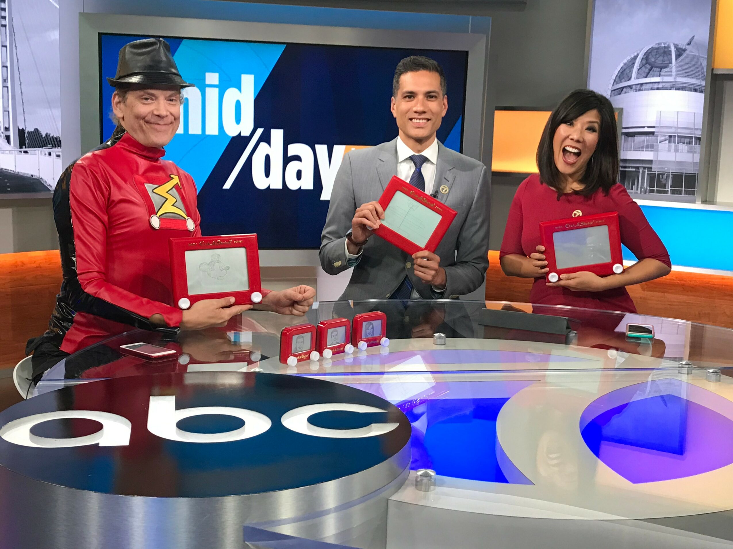 Three news anchors holding tablets and smiling in a television studio with ABC logo, colorful background, and a news set.