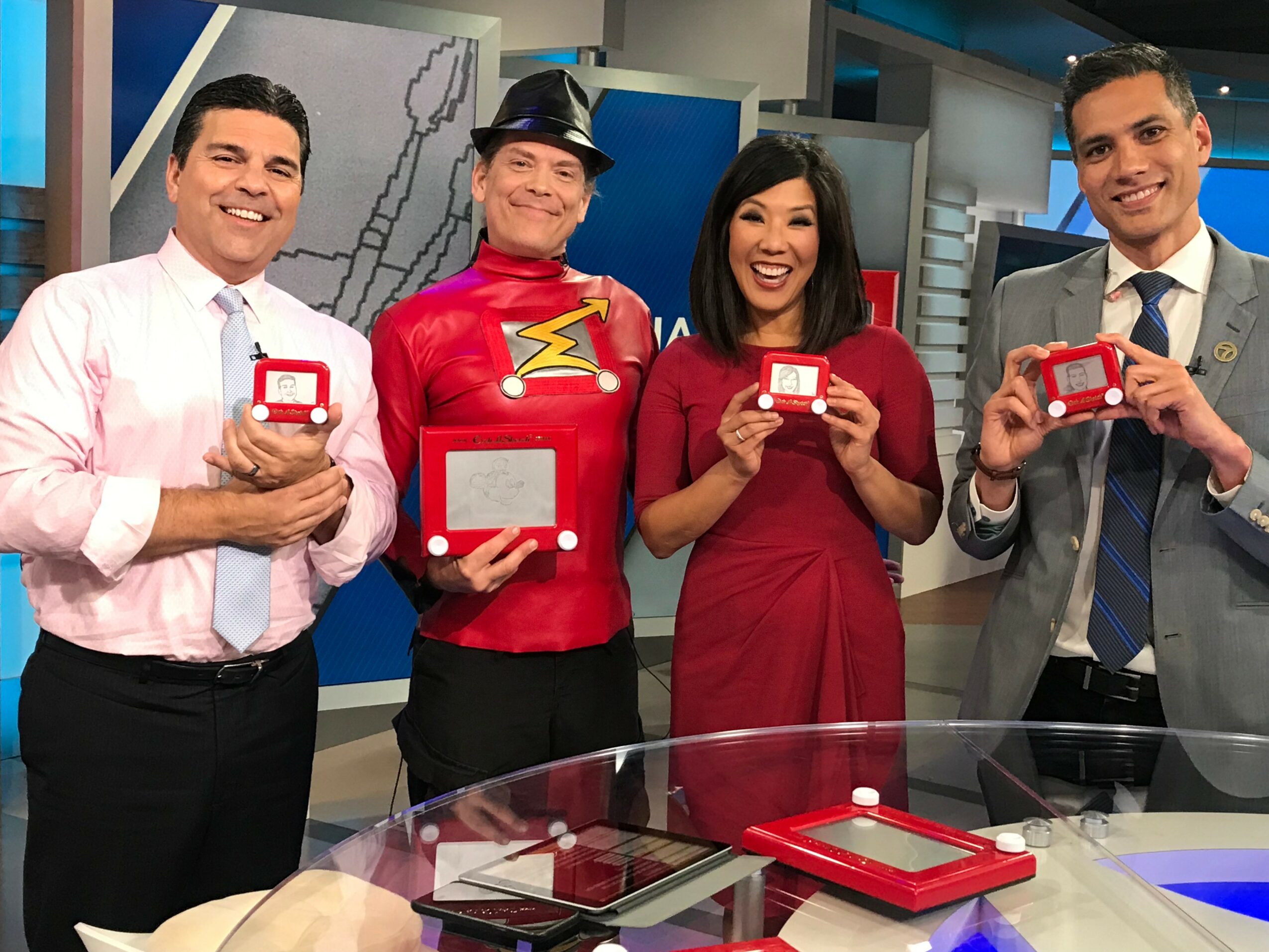 Four news anchors smiling and holding red devices in a studio with a glass table and background screens.