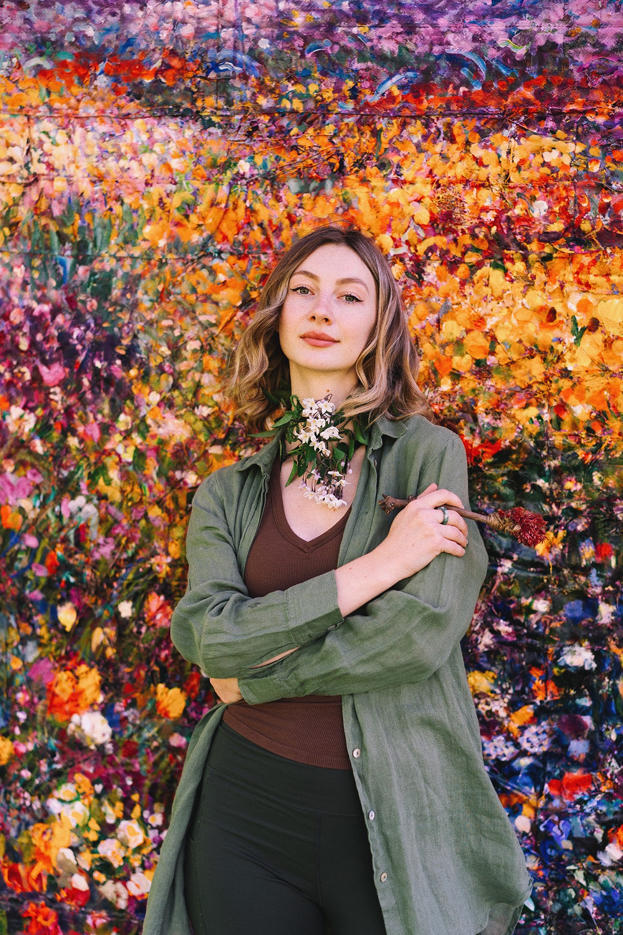 Young woman with shoulder-length hair stands in front of a colorful flower field, wearing a green jacket and brown top.