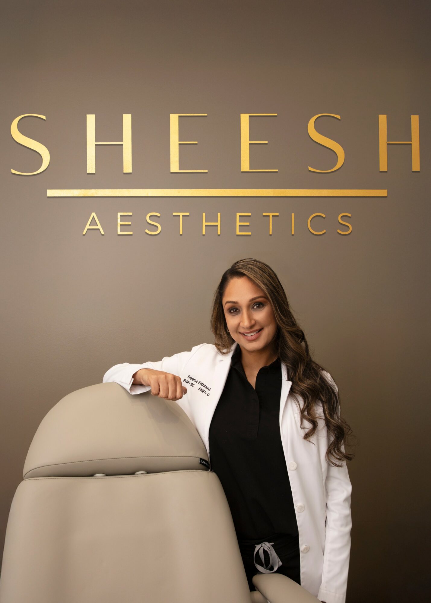 A woman in a white coat standing next to a medical device, smiling, in front of a wall with the words 'SHESH AESTHETICS'.