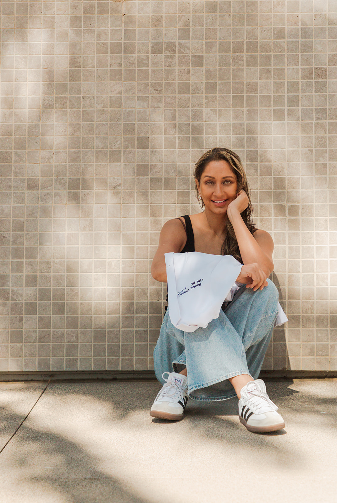 Woman sitting on the ground, smiling, against a tiled wall, wearing a black top, light blue jeans, and white sneakers.