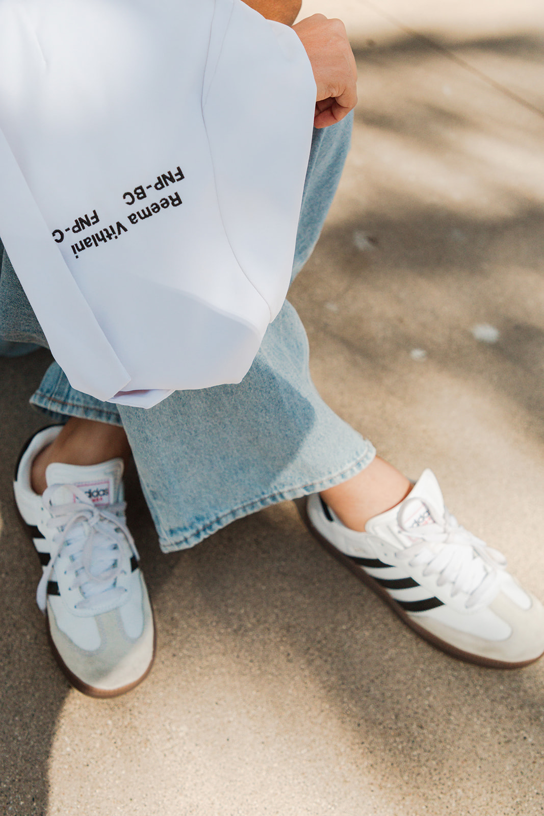 Person sitting outdoors with legs crossed, wearing jeans and white sneakers, holding a white shopping bag with black text.
