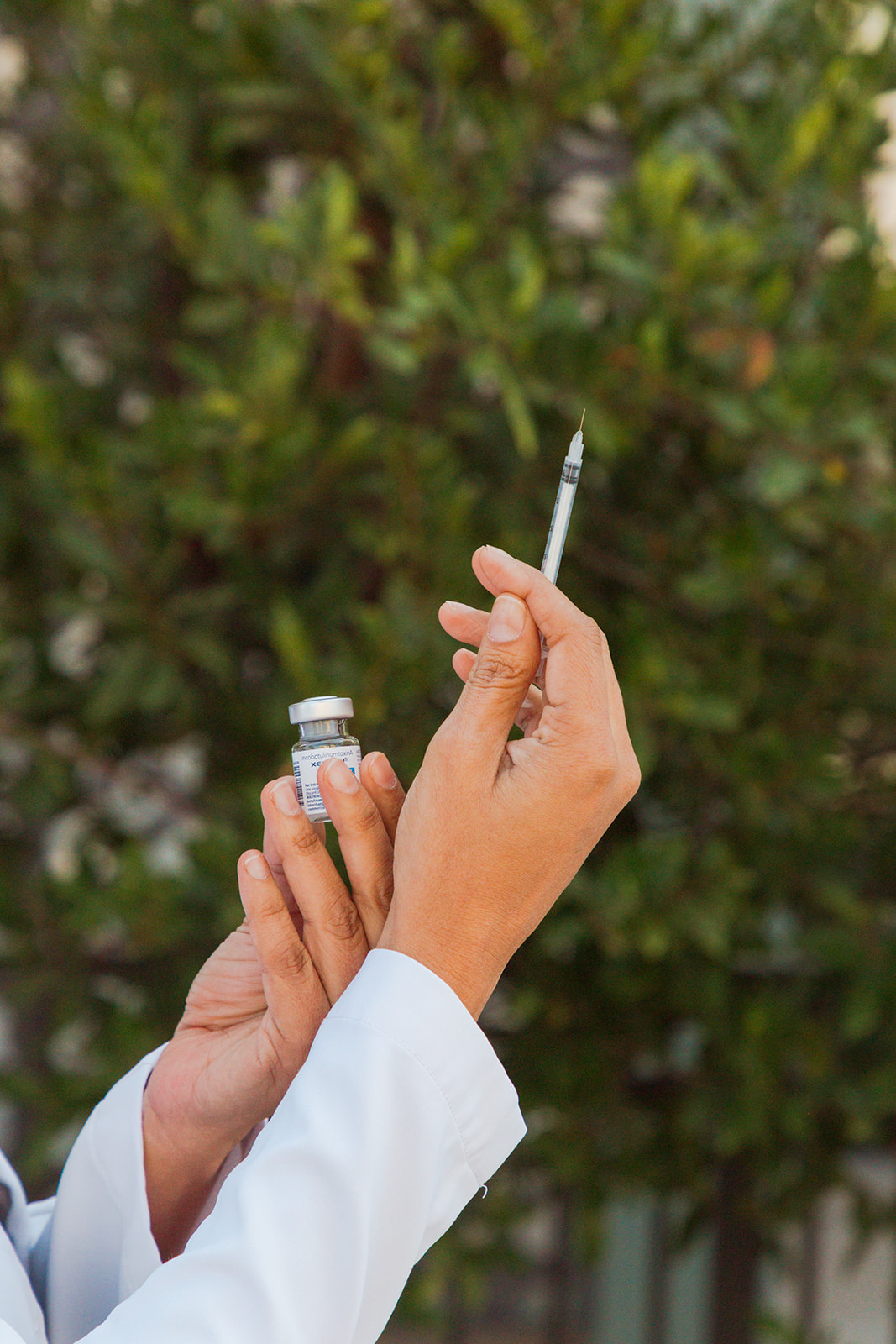 Hand holding a syringe and vial against a green leafy background, person wearing a white coat.