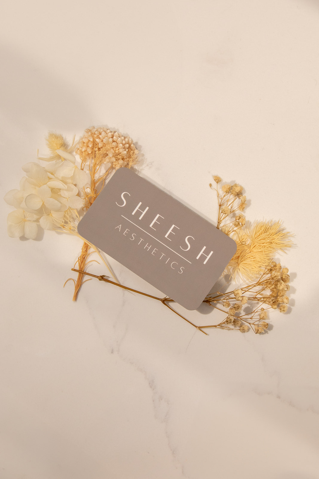 Gray rectangular box labeled 'SHEESH AESTHETICS' surrounded by dried flowers on a white surface.