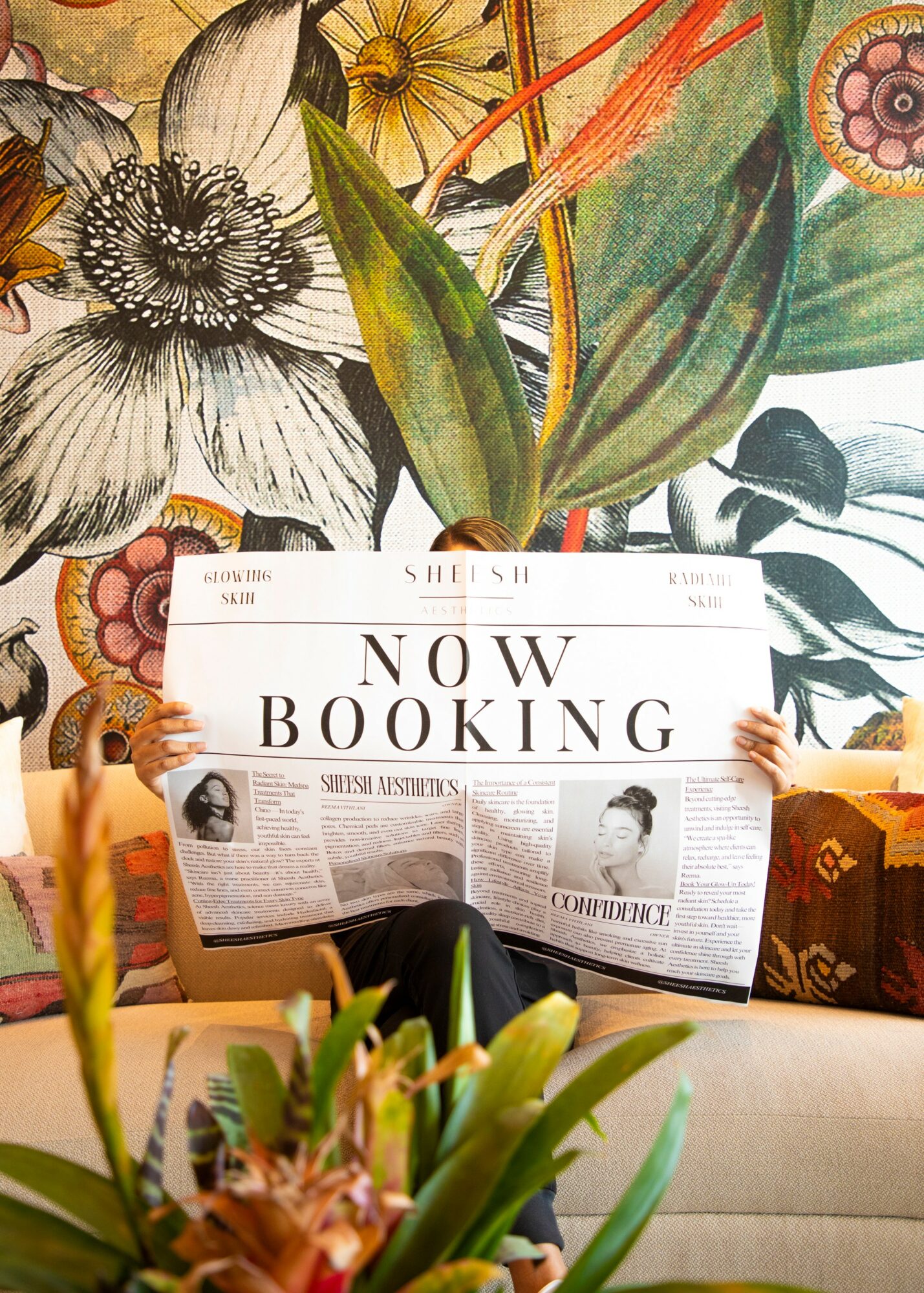 Person holding a newspaper with 'Now Booking' headline, sitting on a sofa with a floral mural background.