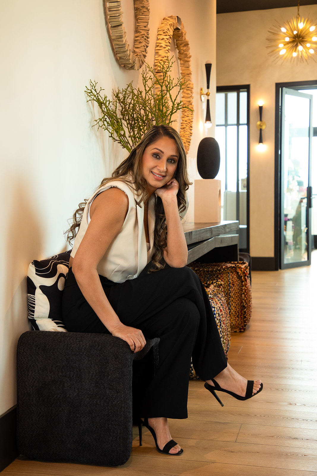Woman sitting on a black sofa, smiling, in a modern interior with plants and decorative items, wooden floor, and glass door.