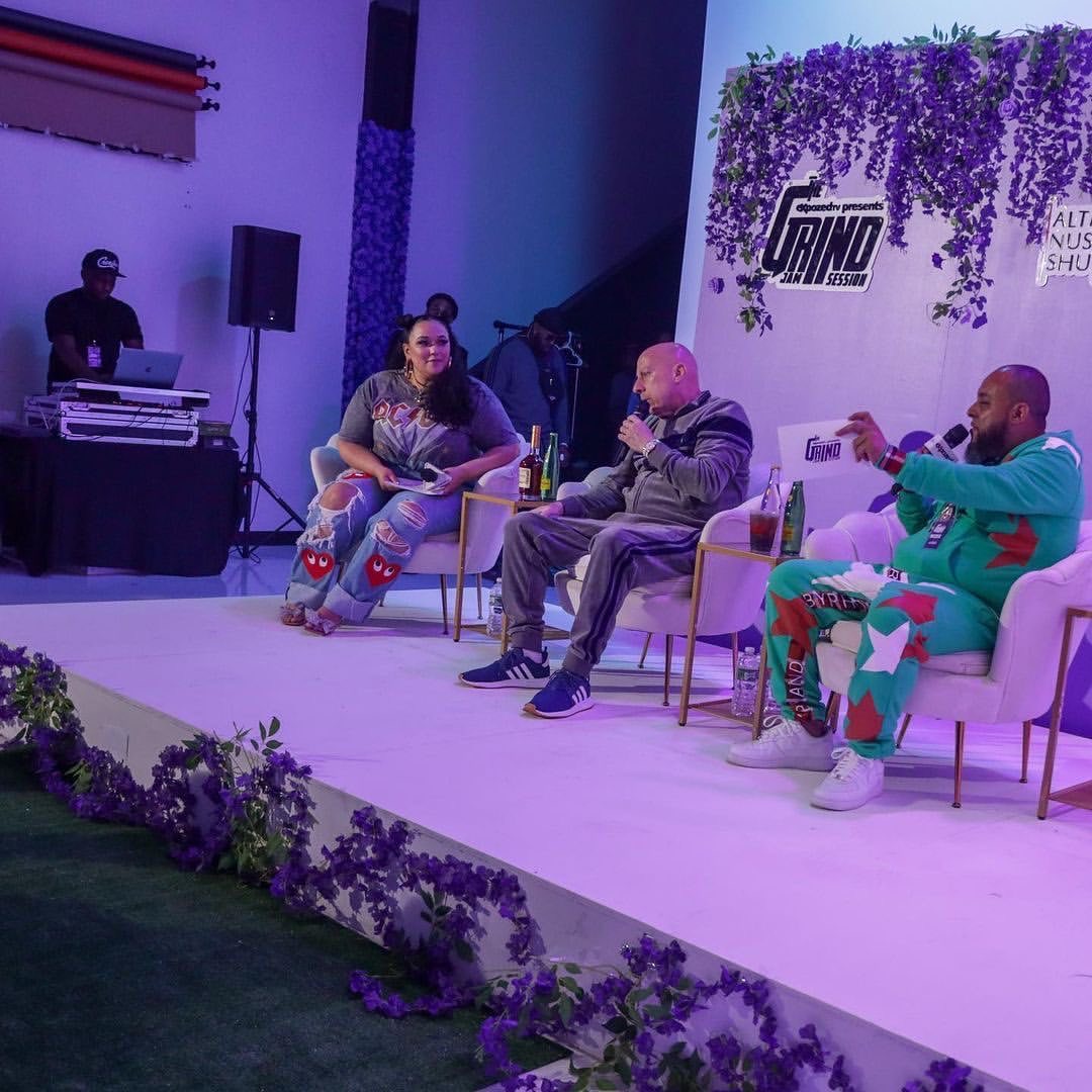 Three people seated on stage with purple floral decorations and a DJ in the background, engaged in conversation.