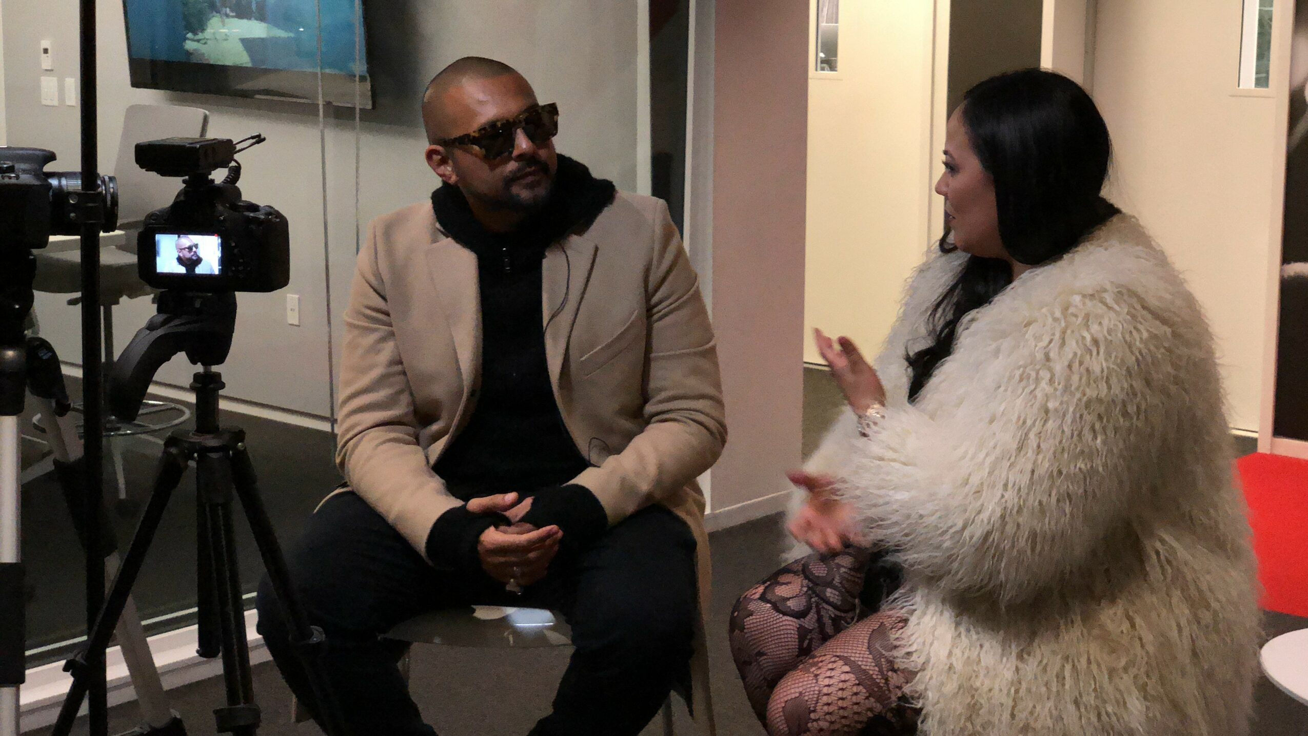 Two people sitting and talking indoors, one with sunglasses and a beige jacket, the other with a white fur coat.