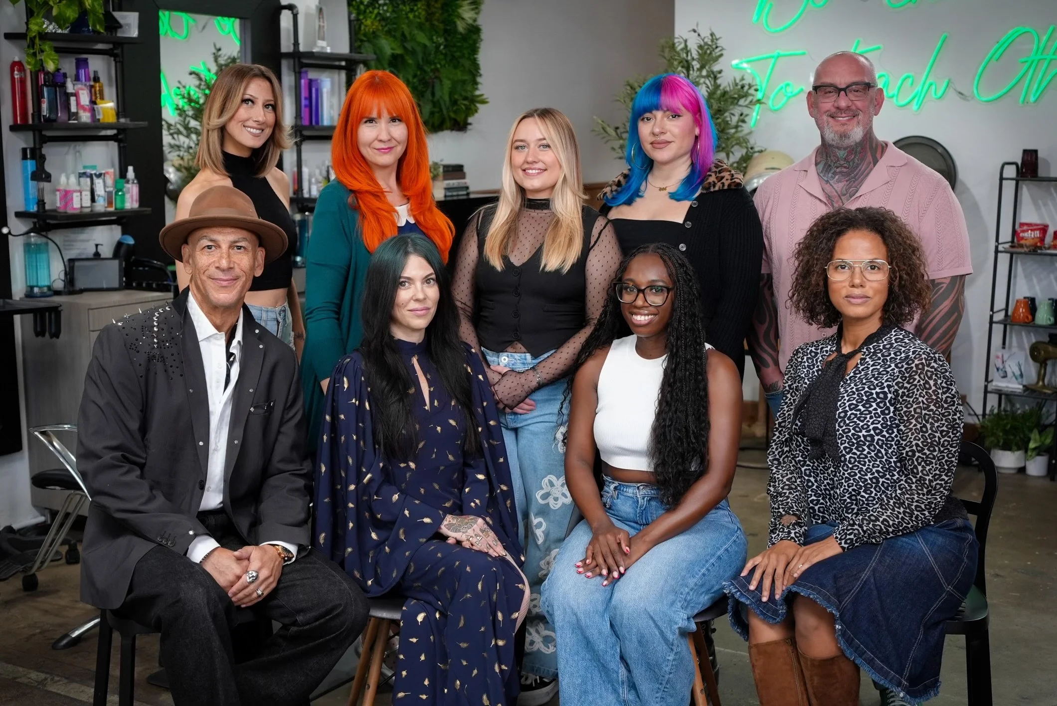 Group of nine diverse people posing in a hair salon, some with colorful hairstyles, smiling and standing or sitting.