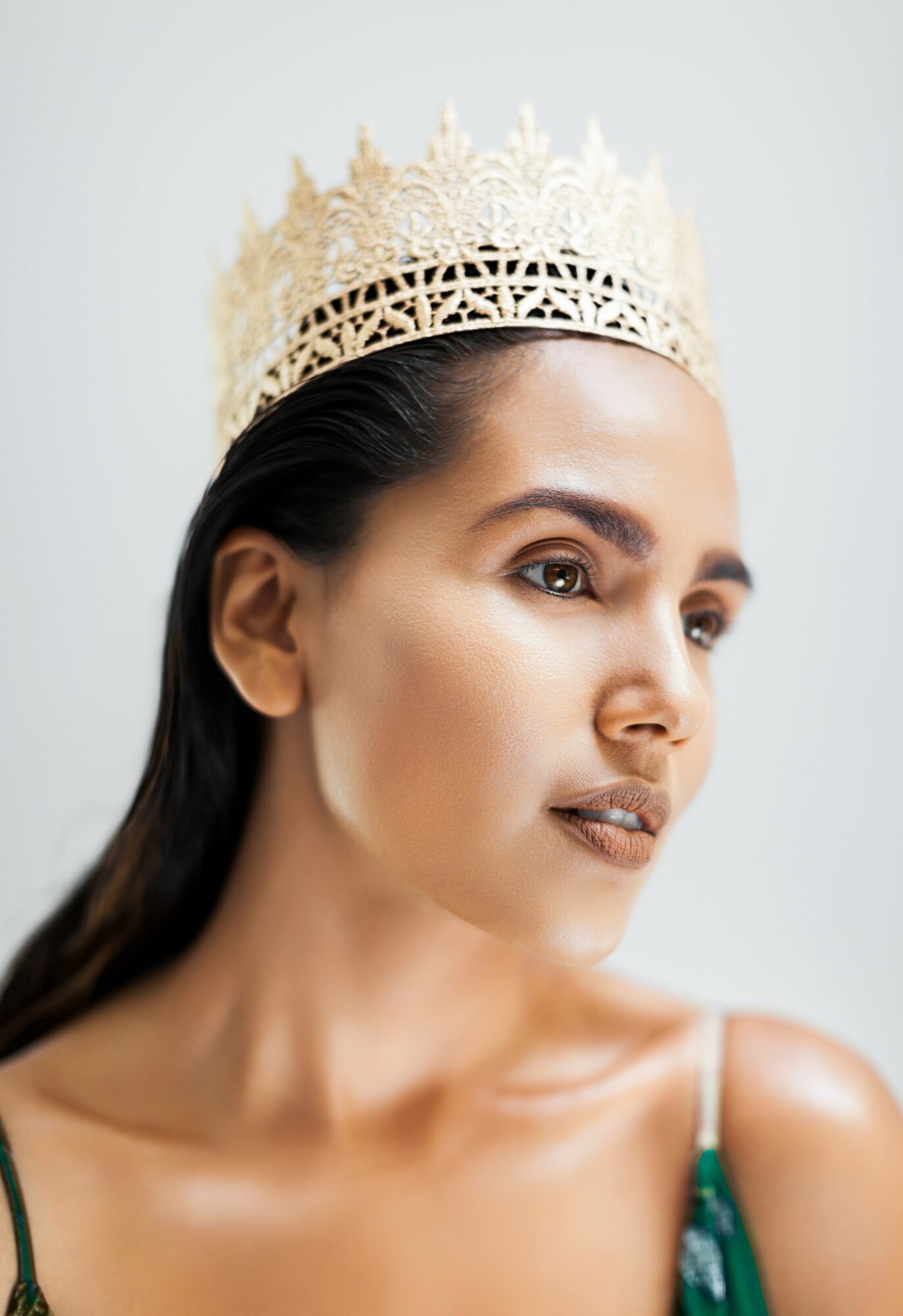 Woman wearing a crown, looking to the side, with dark hair and a strap top, against a plain background.