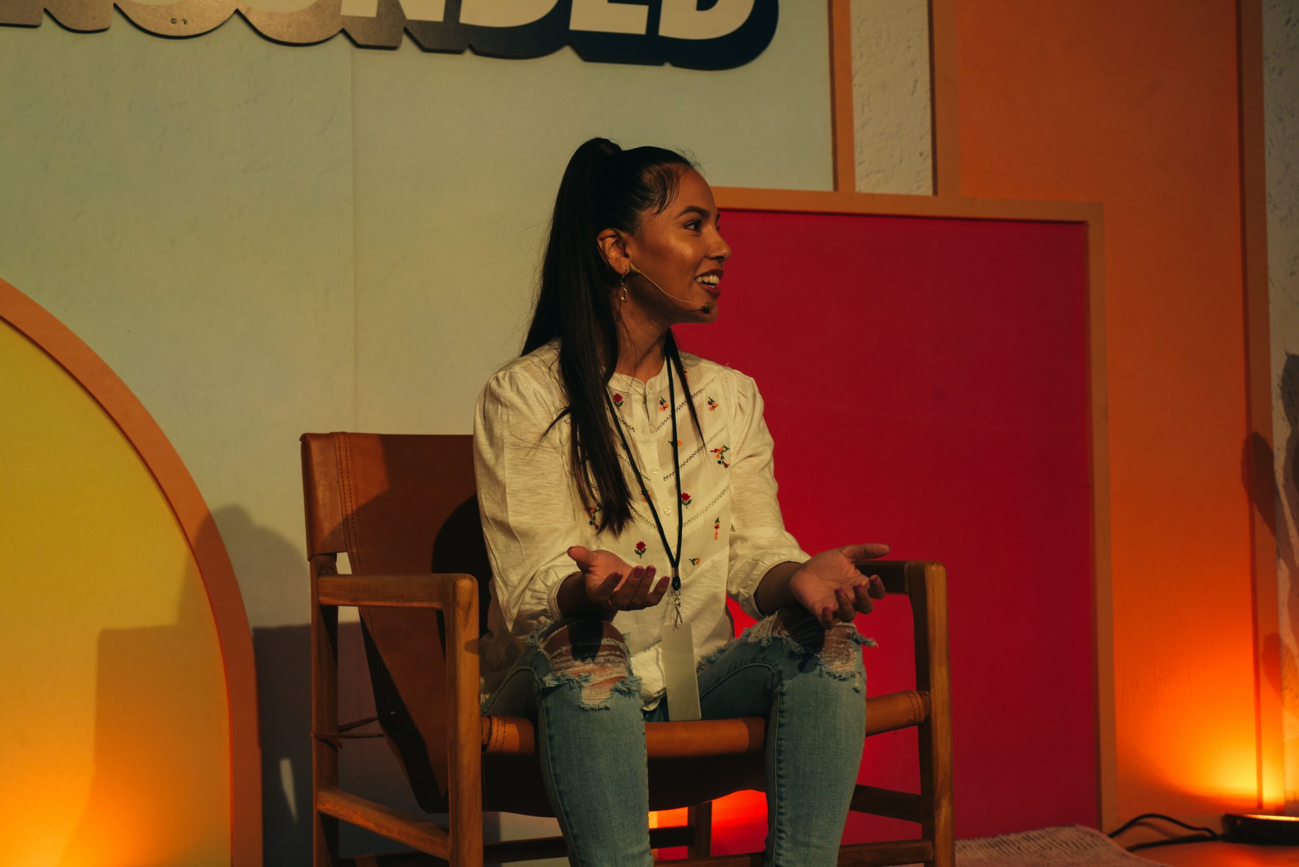 Woman sitting on a wooden chair, gesturing with hands, in a brightly lit room with colorful background.