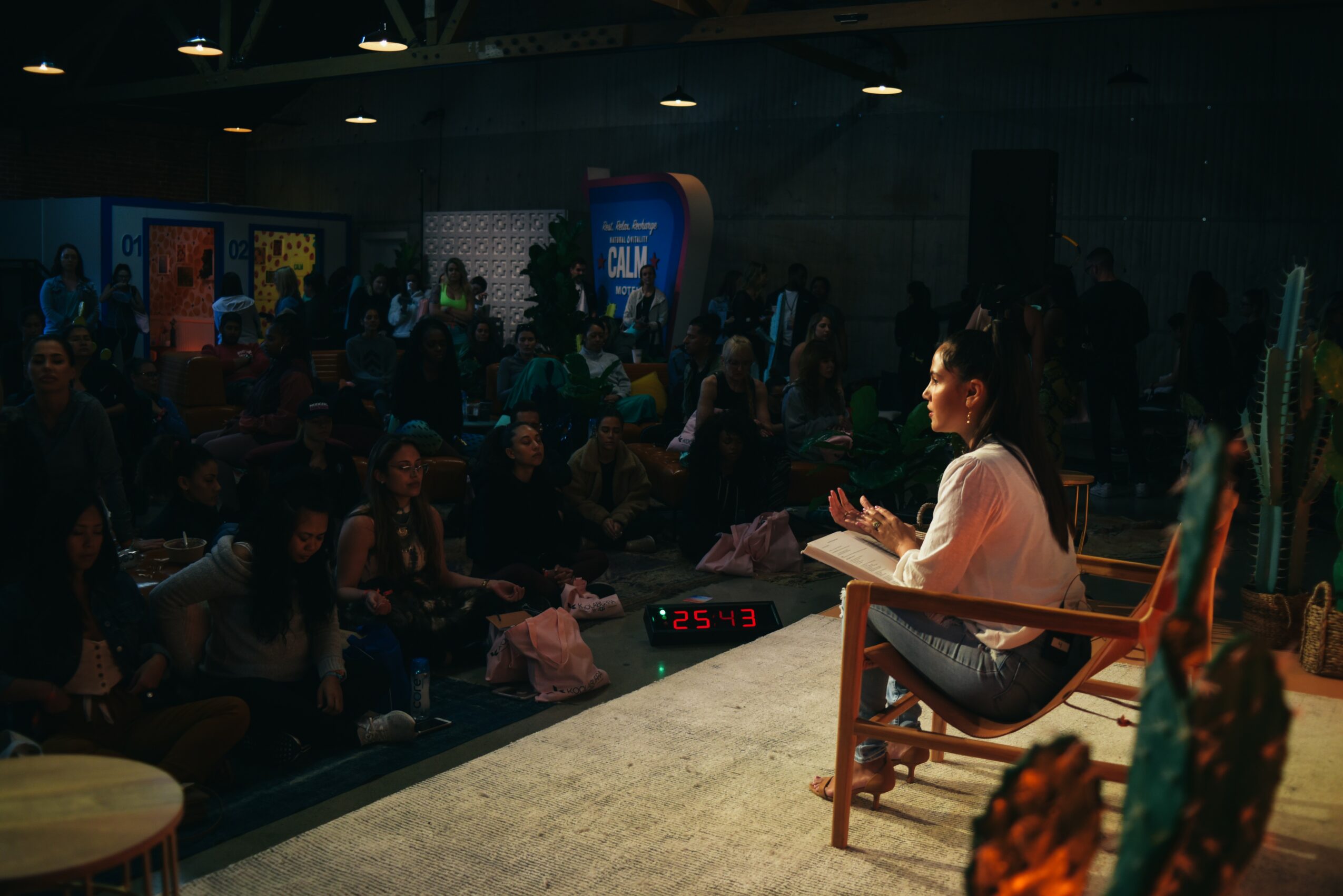 Woman sitting on a chair on stage, speaking to an audience in a large indoor space, with banners and people in the background.