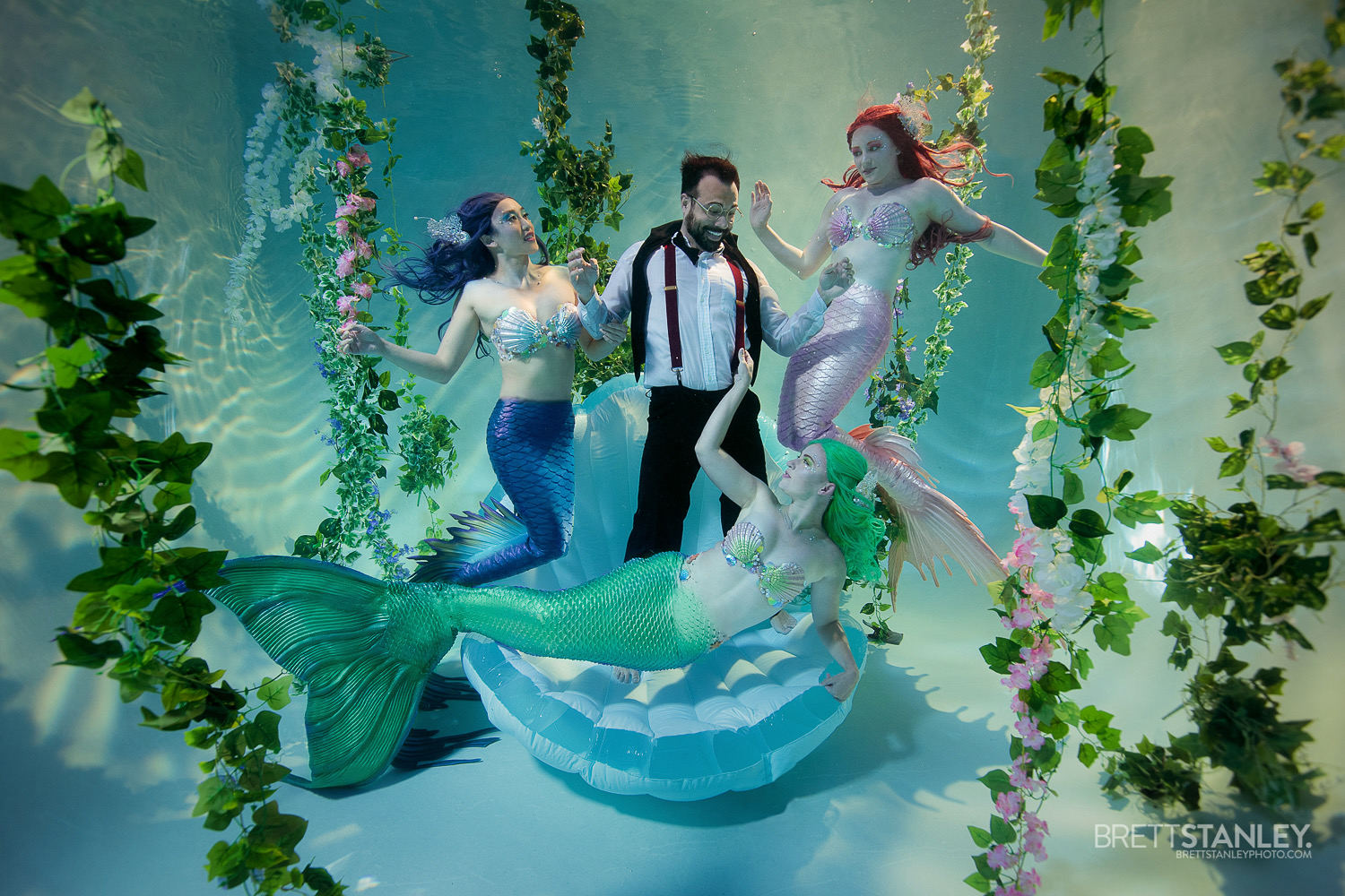 Three mermaids and a person in a tuxedo pose underwater surrounded by green plants and pink flowers, with a mermaid tail on a platform.