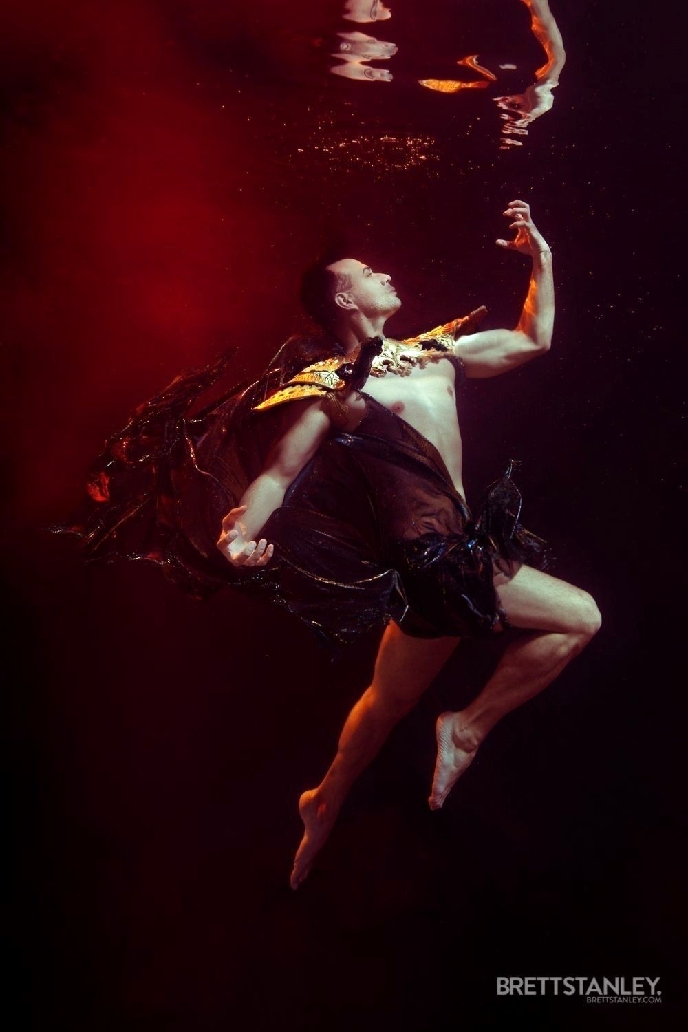 Person underwater with head tilted back, arm raised, and hair flowing, in a dark environment with red lighting.