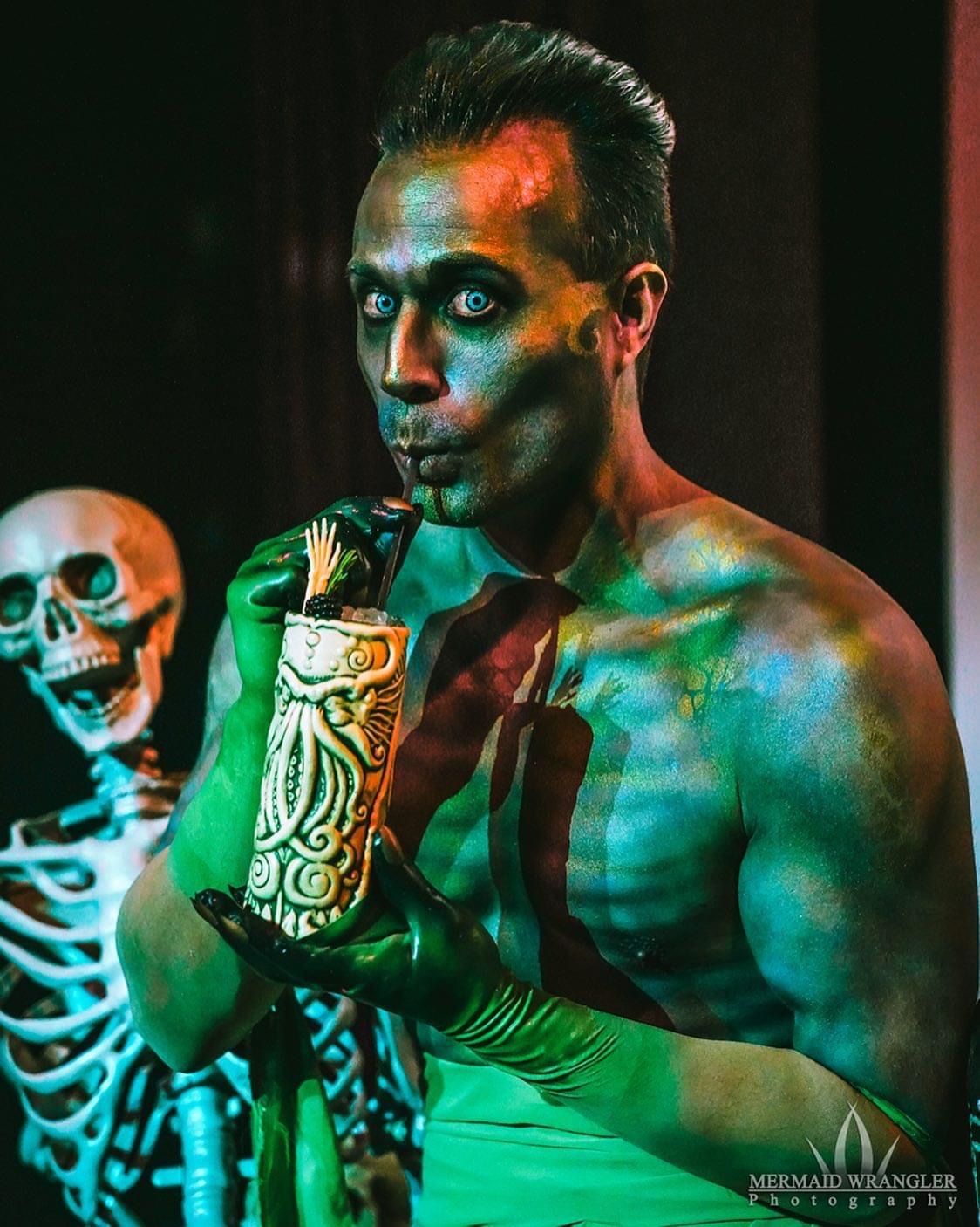 Person with makeup and costume, holding a decorated object, with a skeleton in the background, in a dark setting.
