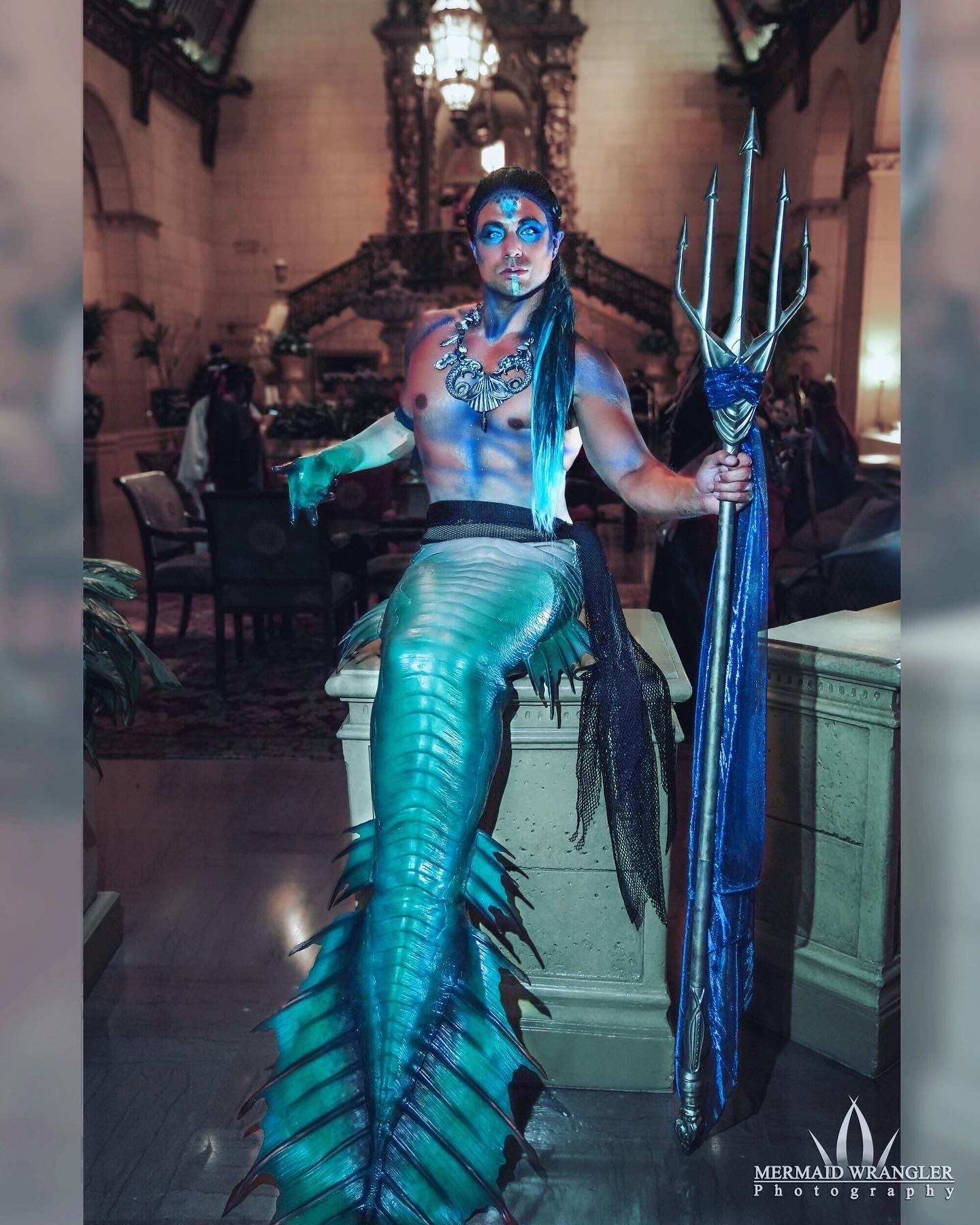 Person dressed as a mermaid with a blue tail, holding a trident, in an ornate indoor setting.
