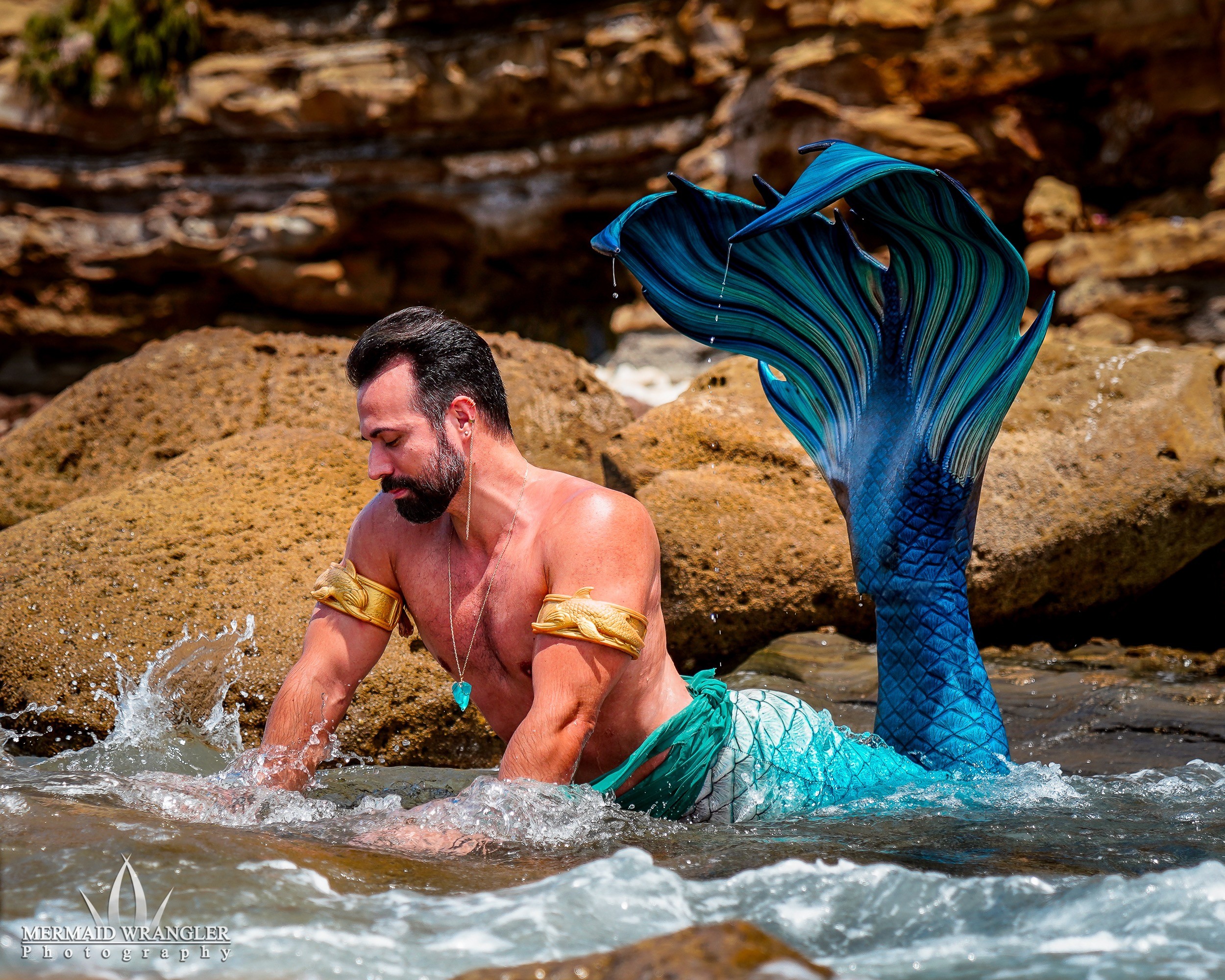 Man with a mermaid tail costume in water near rocks, smiling and splashing water, outdoors.