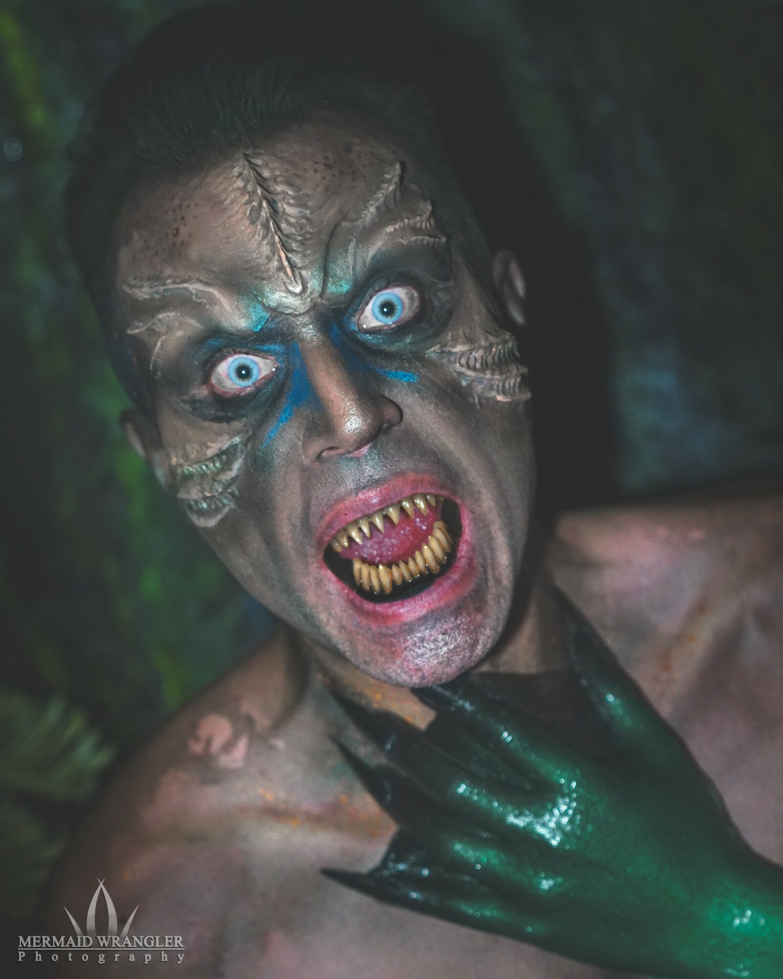 Person with face painted as a monster, mouth open showing sharp teeth, eyes wide, dark background, hand on neck.
