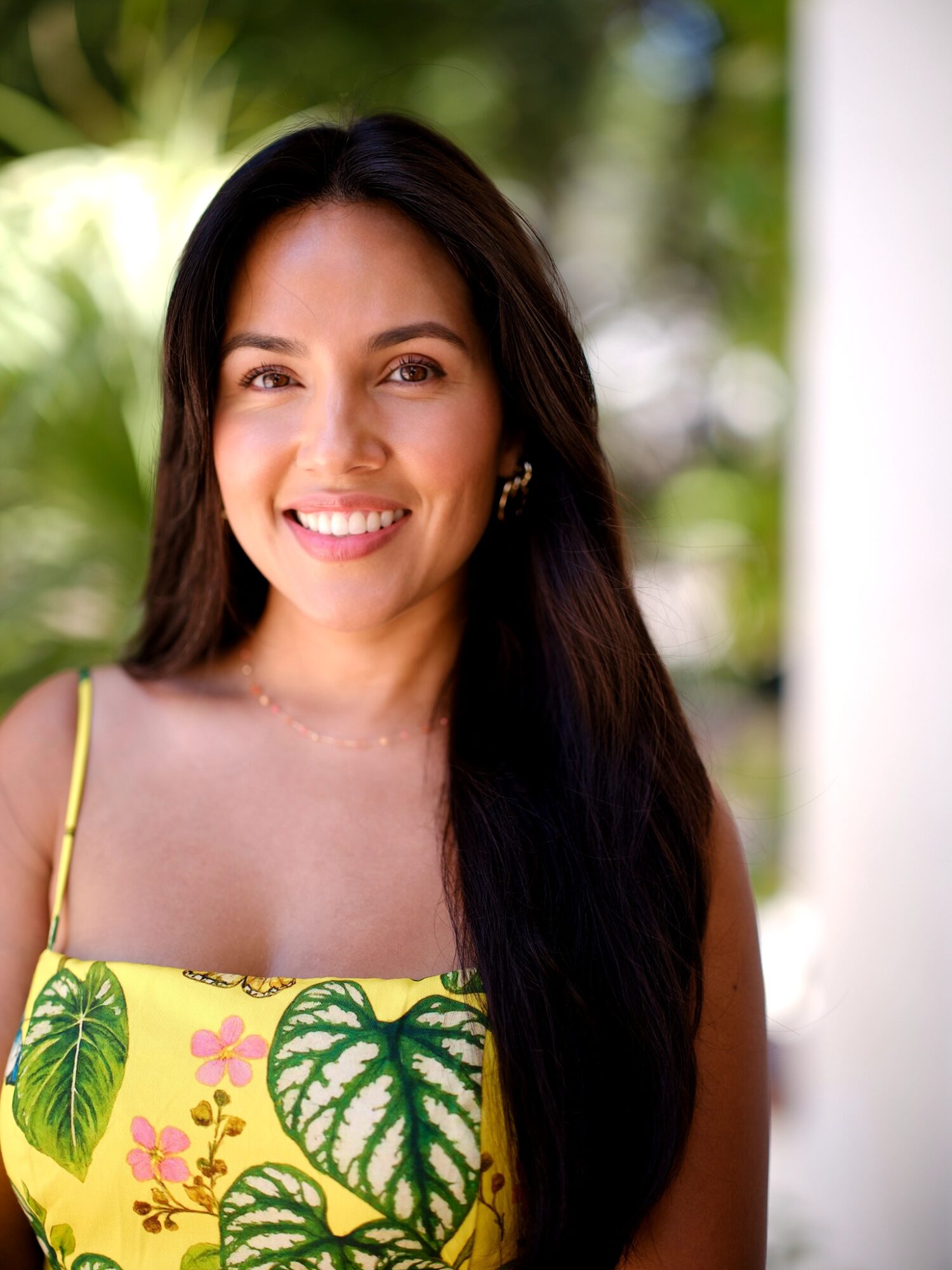 Smiling woman with long dark hair outdoors, wearing a yellow dress with green leaf patterns, background blurred greenery