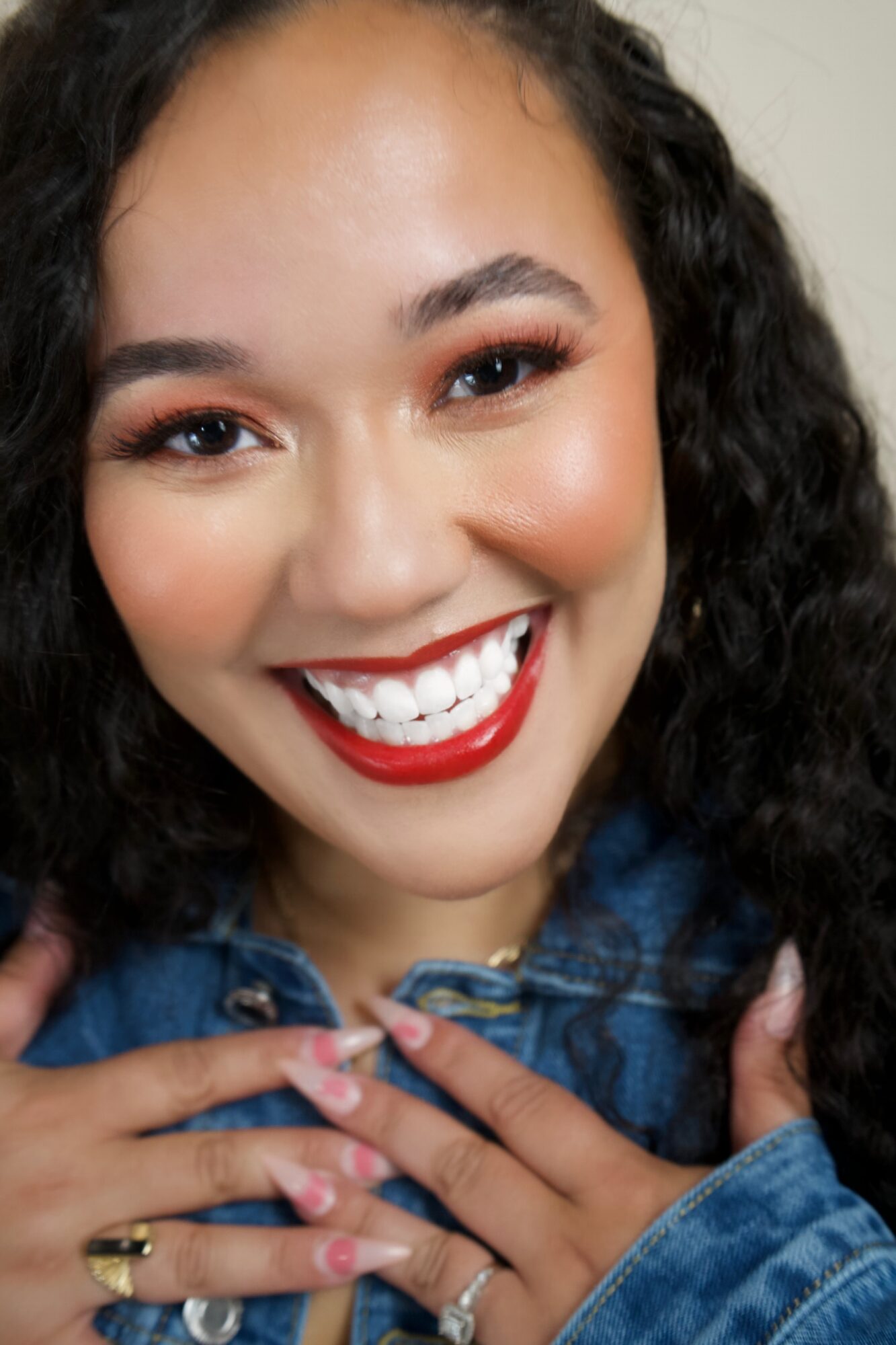 Close-up of smiling woman with curly hair, red lipstick, and hands near her chest, wearing a denim jacket.