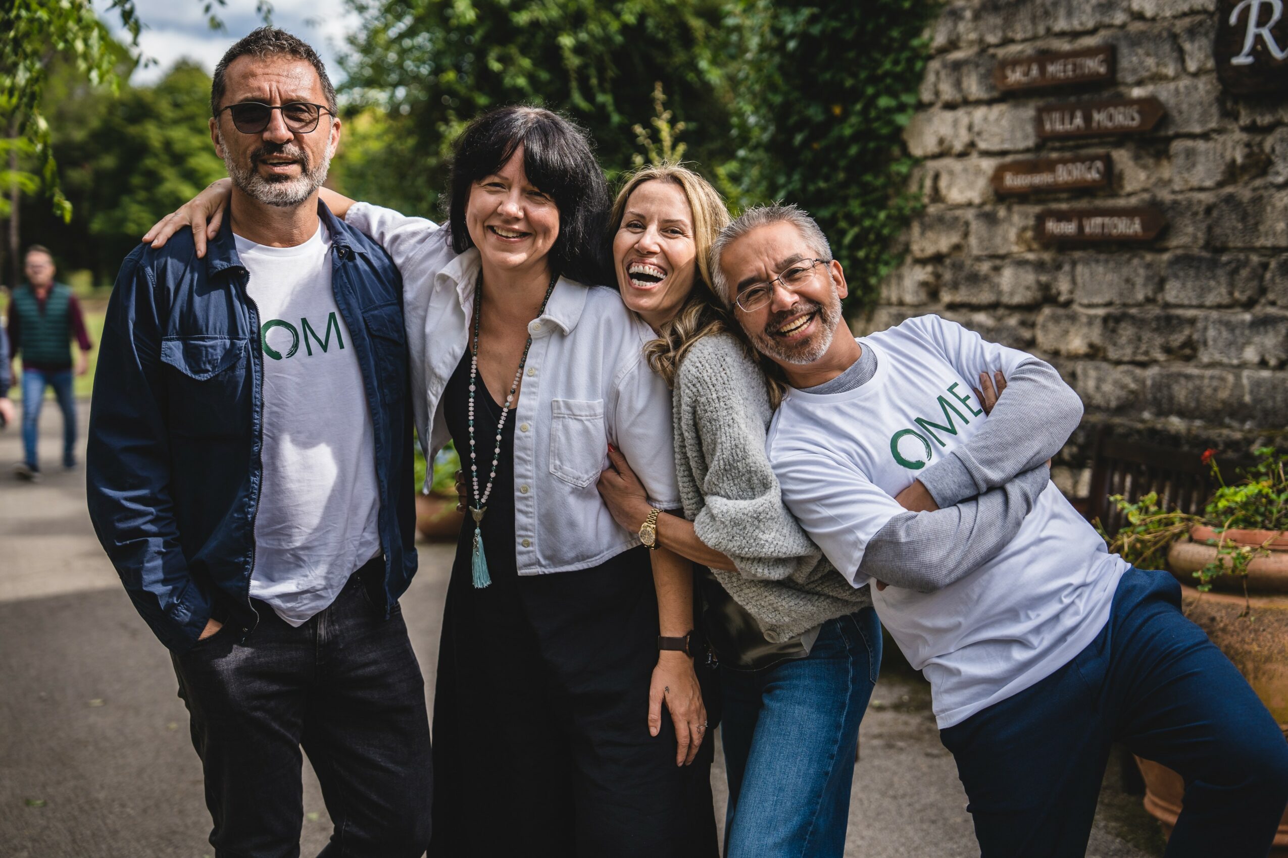 Group of four smiling people standing outdoors, with trees and a stone wall in the background.