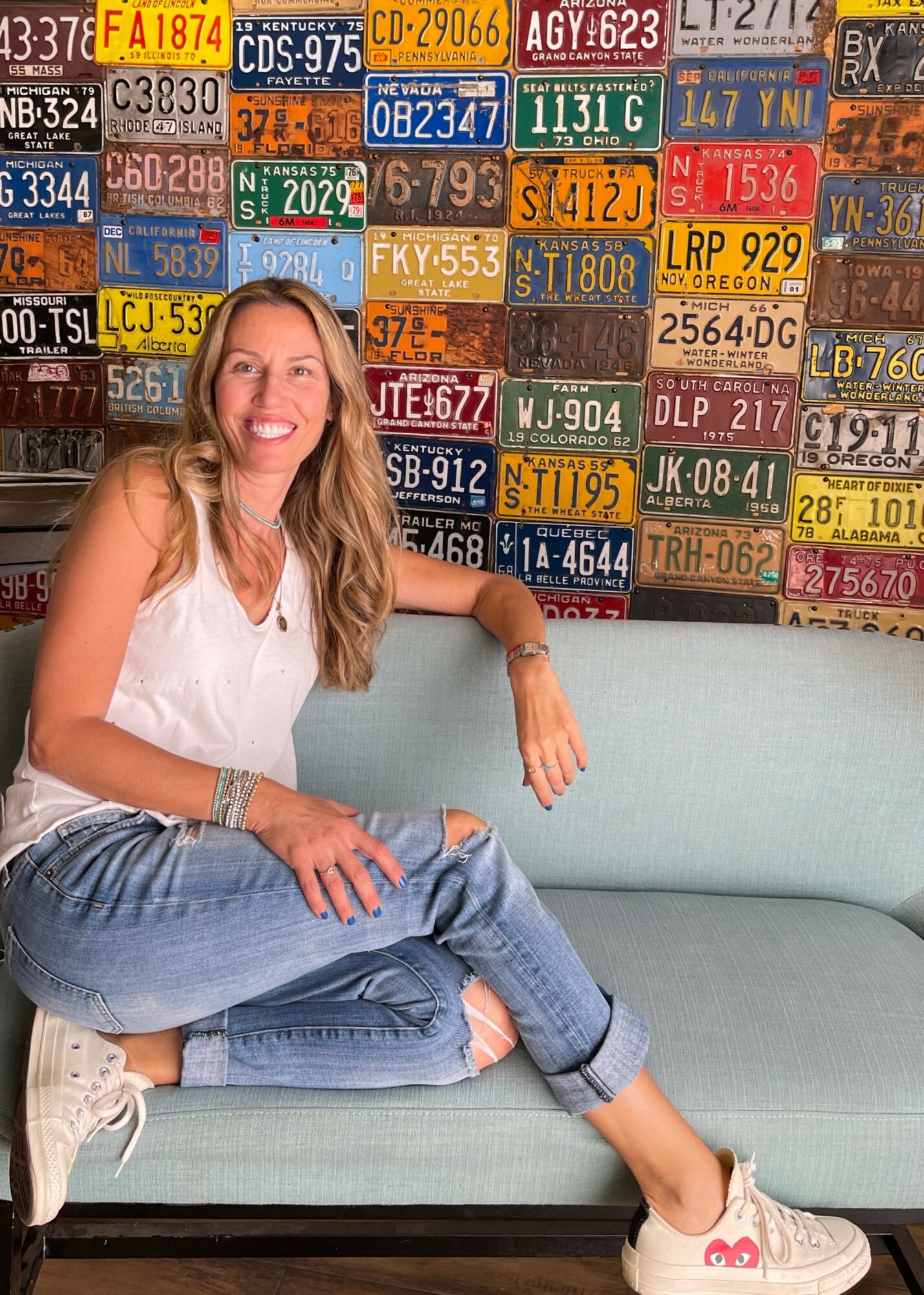 Woman with long hair sitting on a light blue couch, smiling, with a wall of colorful license plates behind her.