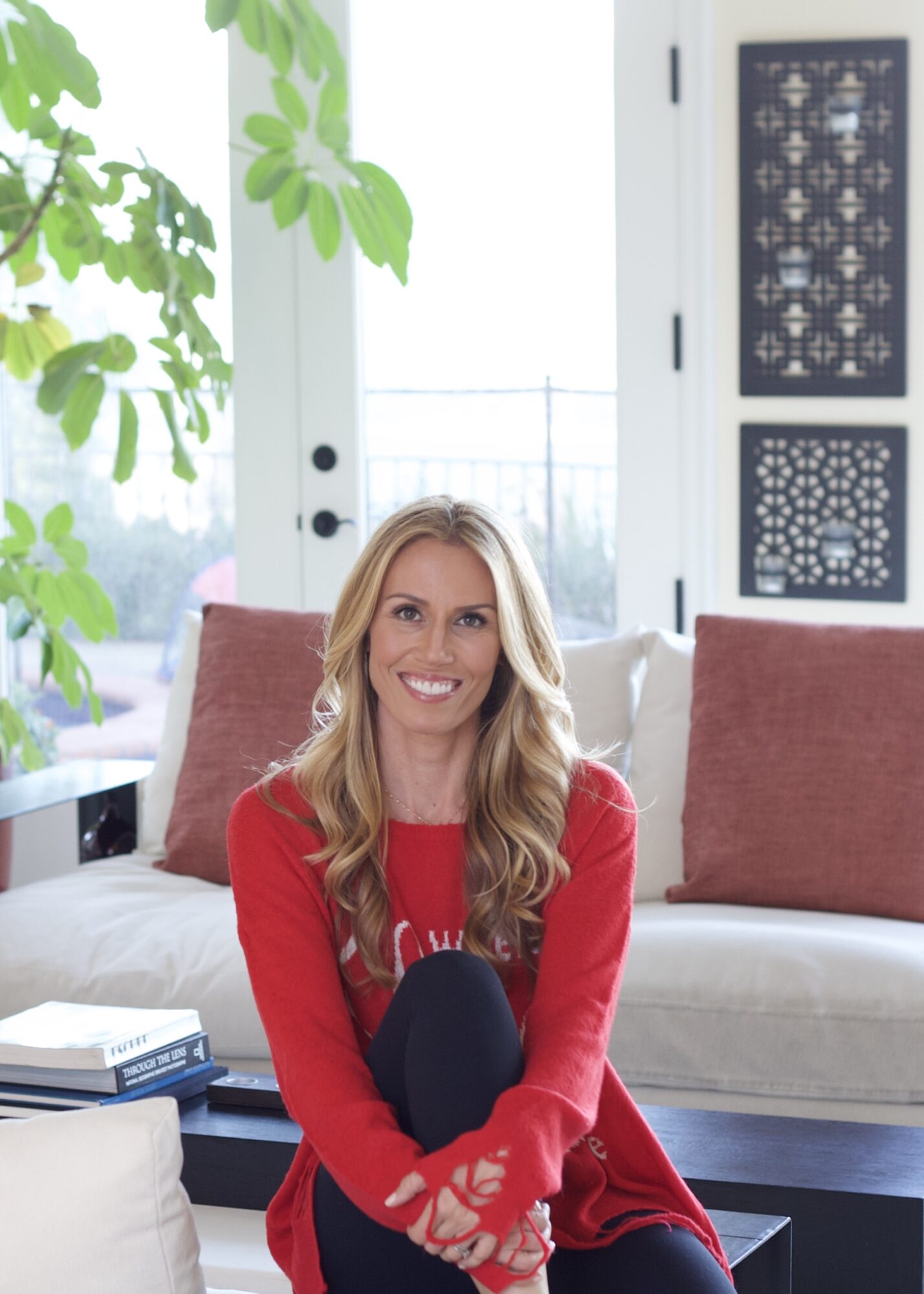 Woman with long blonde hair sitting on a sofa in a bright room, smiling, wearing a red sweater, with plants and decorative panels in the background.