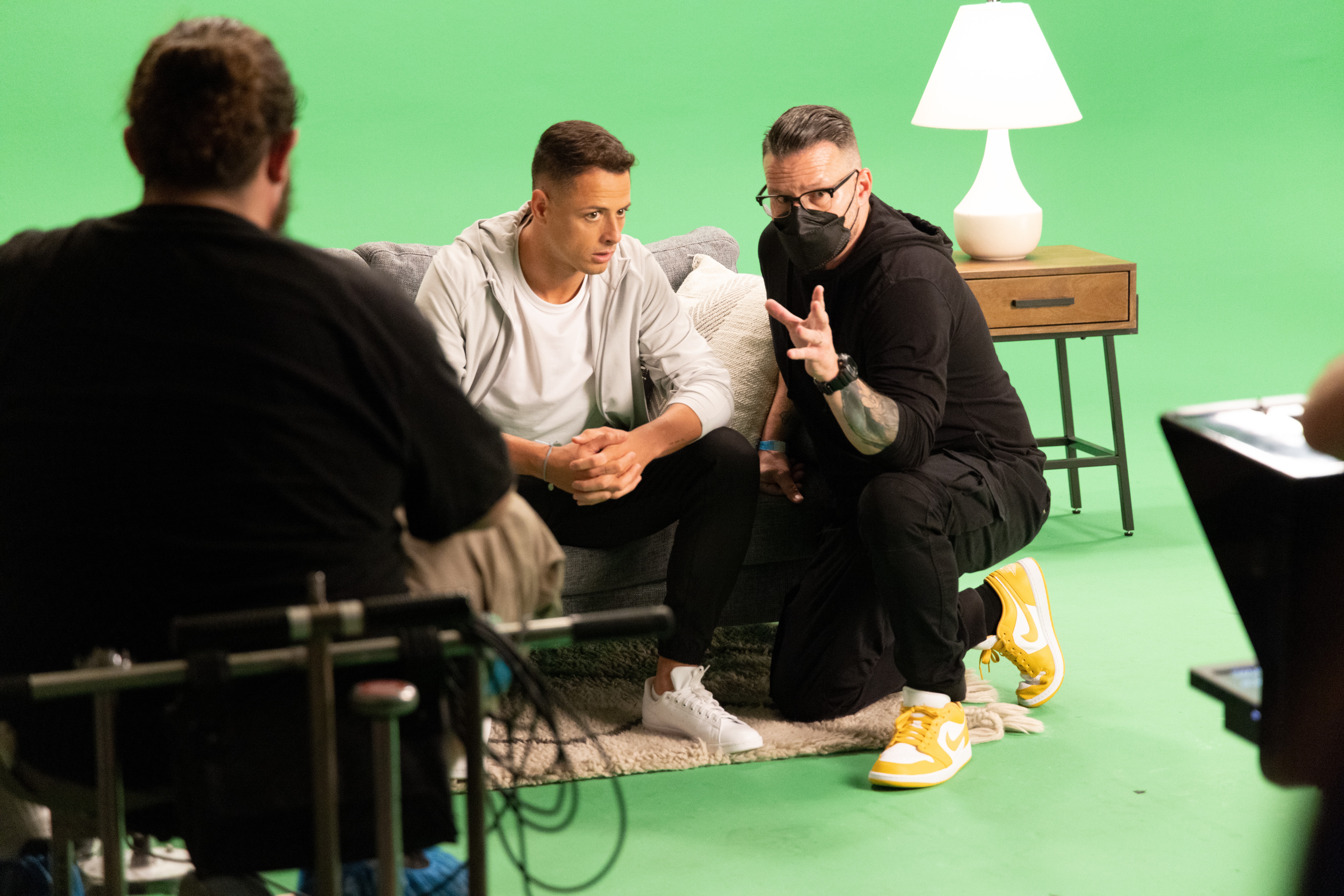Three people in a studio with a green screen, one sitting on a couch, two kneeling, discussing, with a lamp on a side table.