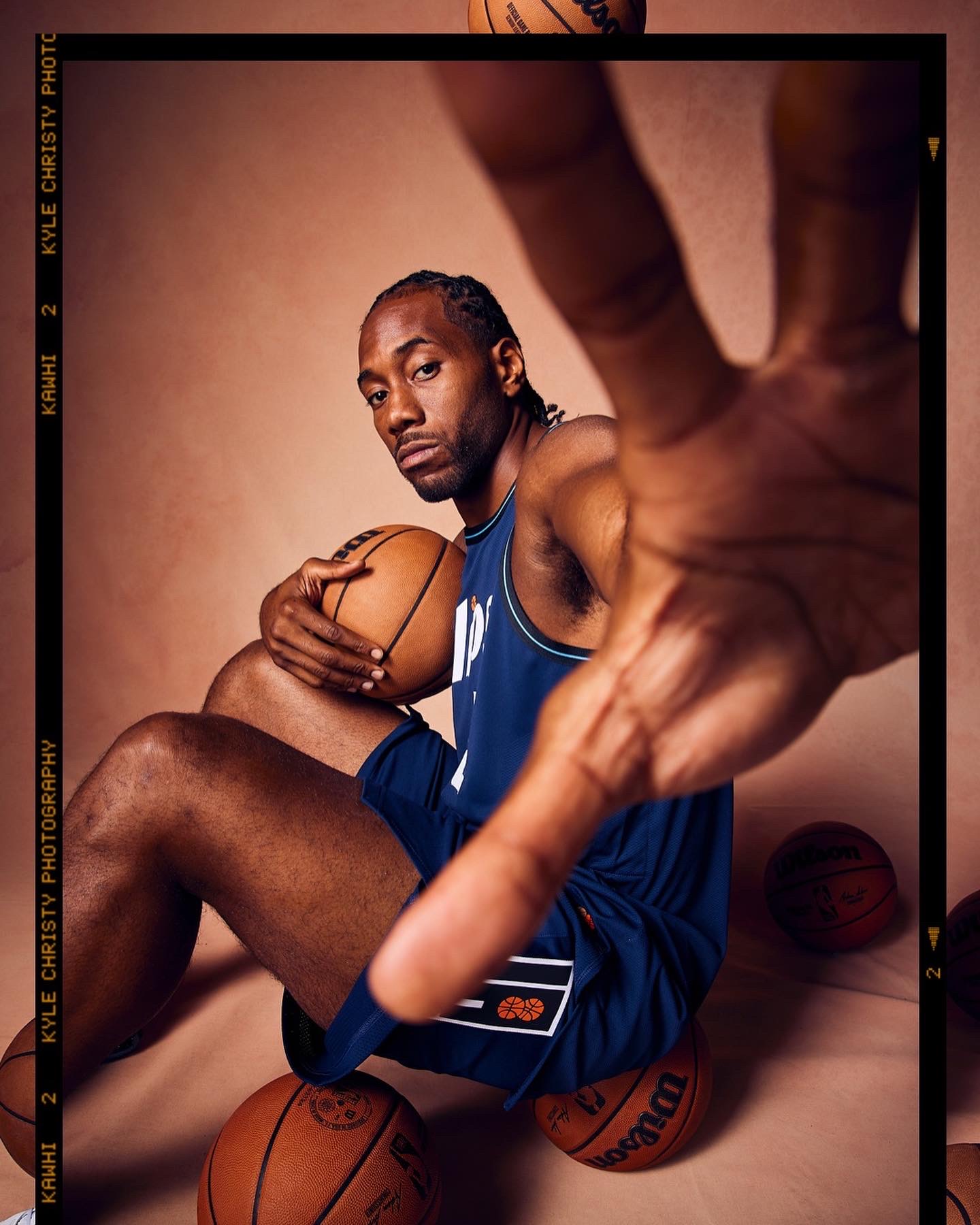 Man in blue sports uniform holding basketball, reaching toward camera with hand, surrounded by basketballs, neutral background.