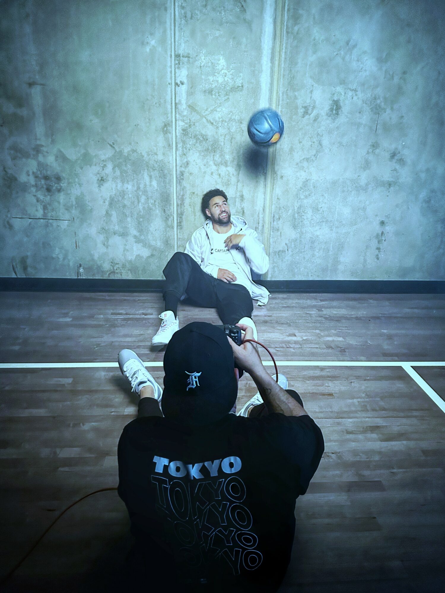 Person sitting on the floor in a gym, with a ball suspended above, and another person taking a photo.