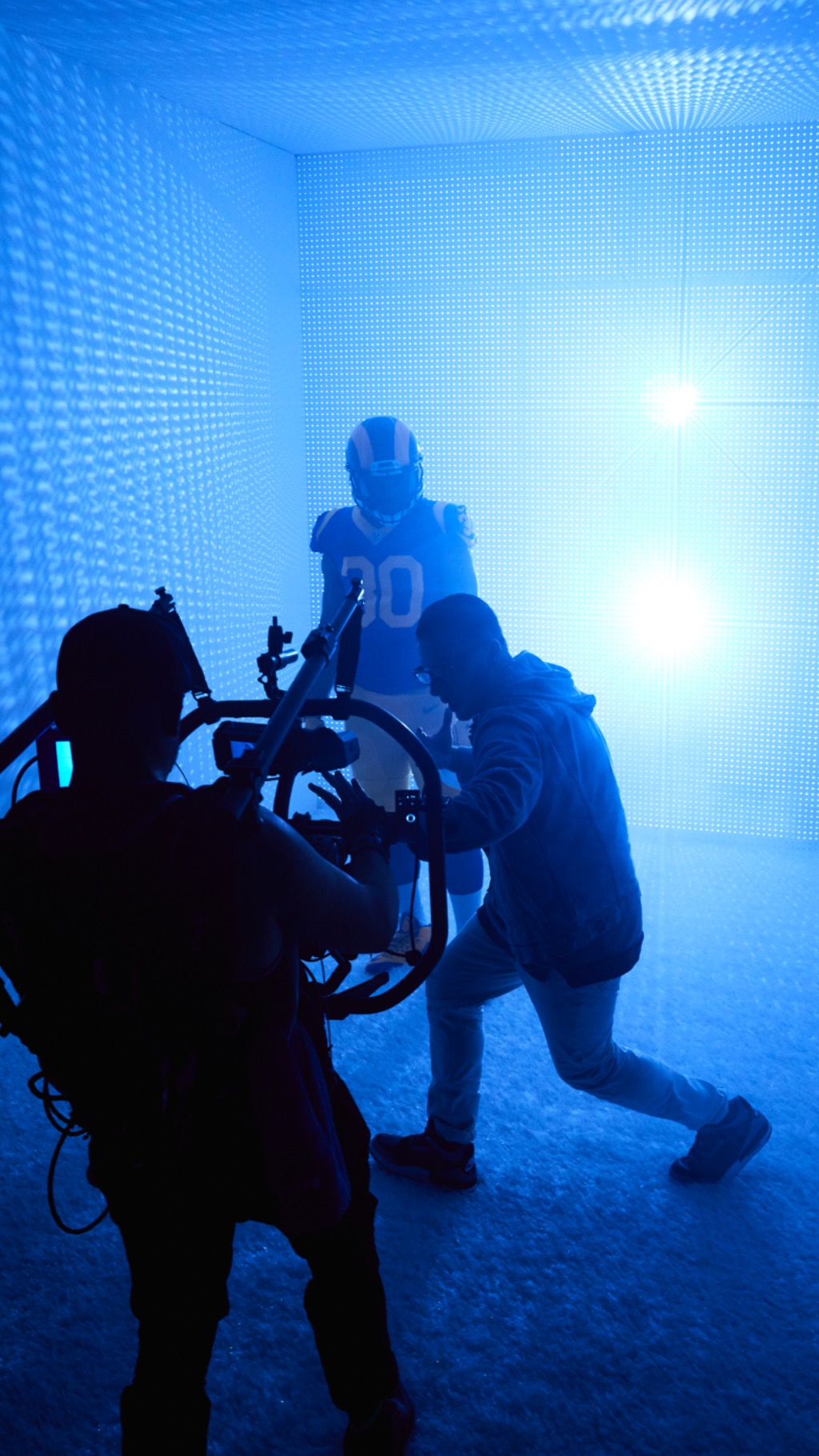 Person in sports jersey kneeling with a person in a helmet and equipment in a blue-lit room.