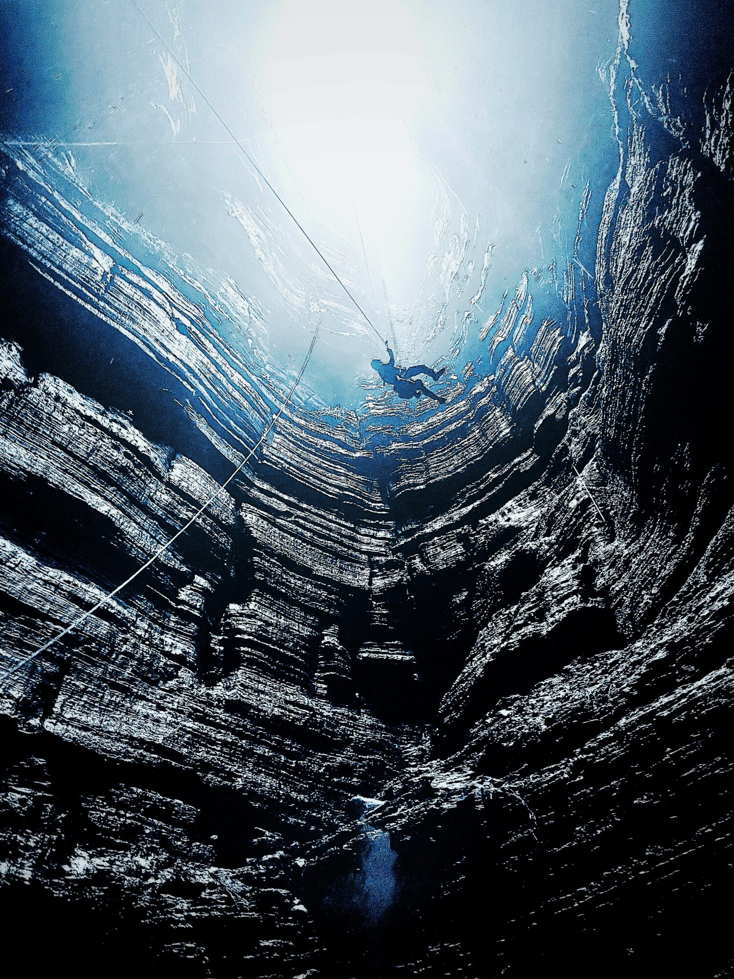 Person rappelling down a deep, narrow canyon with sunlight shining from above, rocky walls on sides, and a rope in hand.