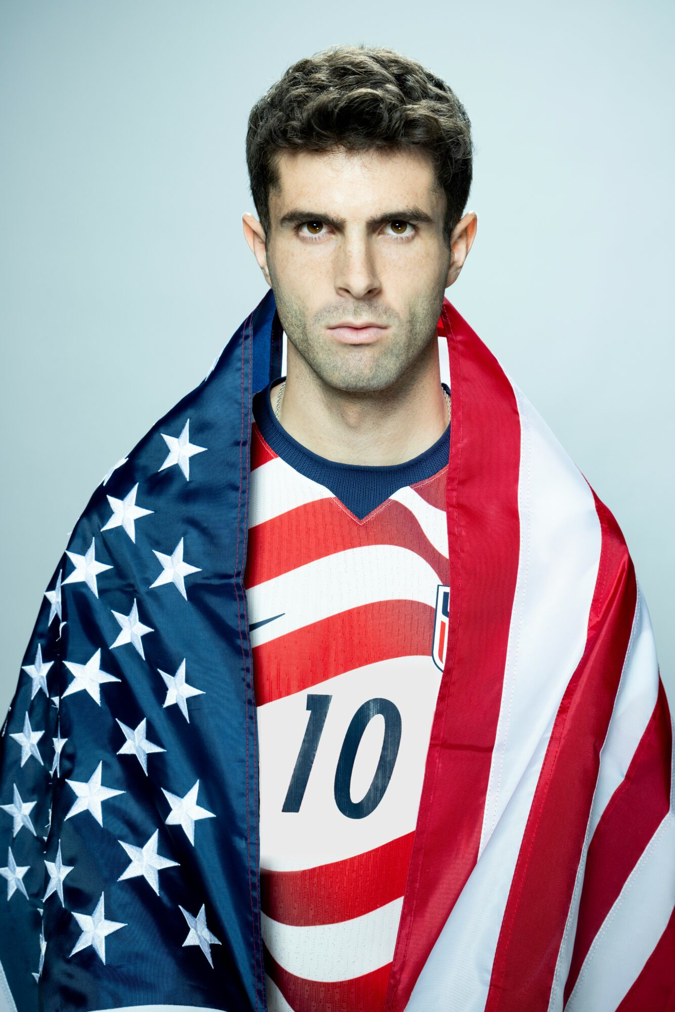 Man with dark hair and serious expression, wearing a sports jersey and draped in an American flag.