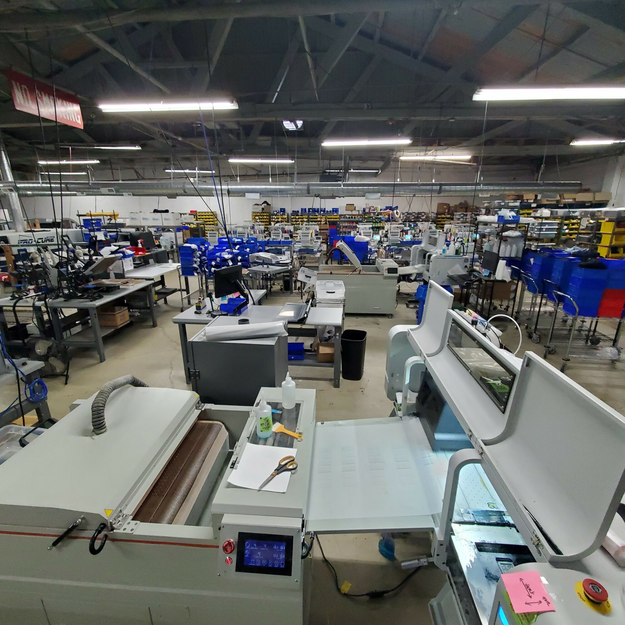 Factory or workshop with machinery and workstations, viewed from a slightly elevated angle, with bright overhead lighting.
