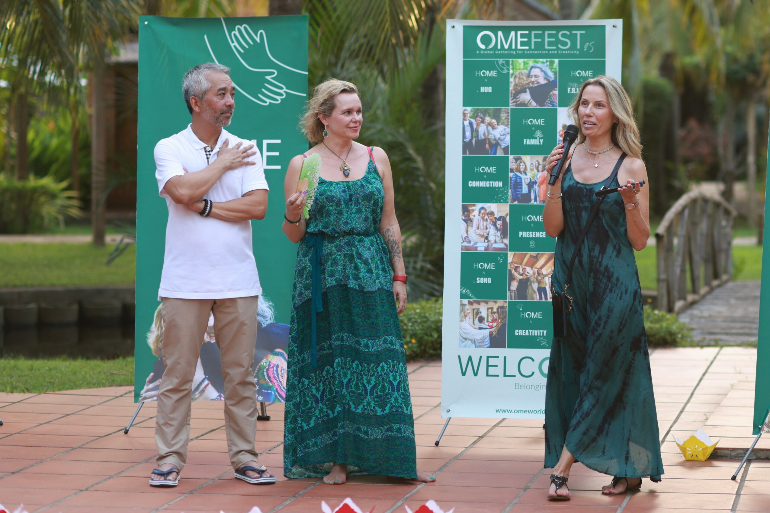 Three people standing outdoors near banners, two women in long dresses and a man in a white shirt, trees in background.